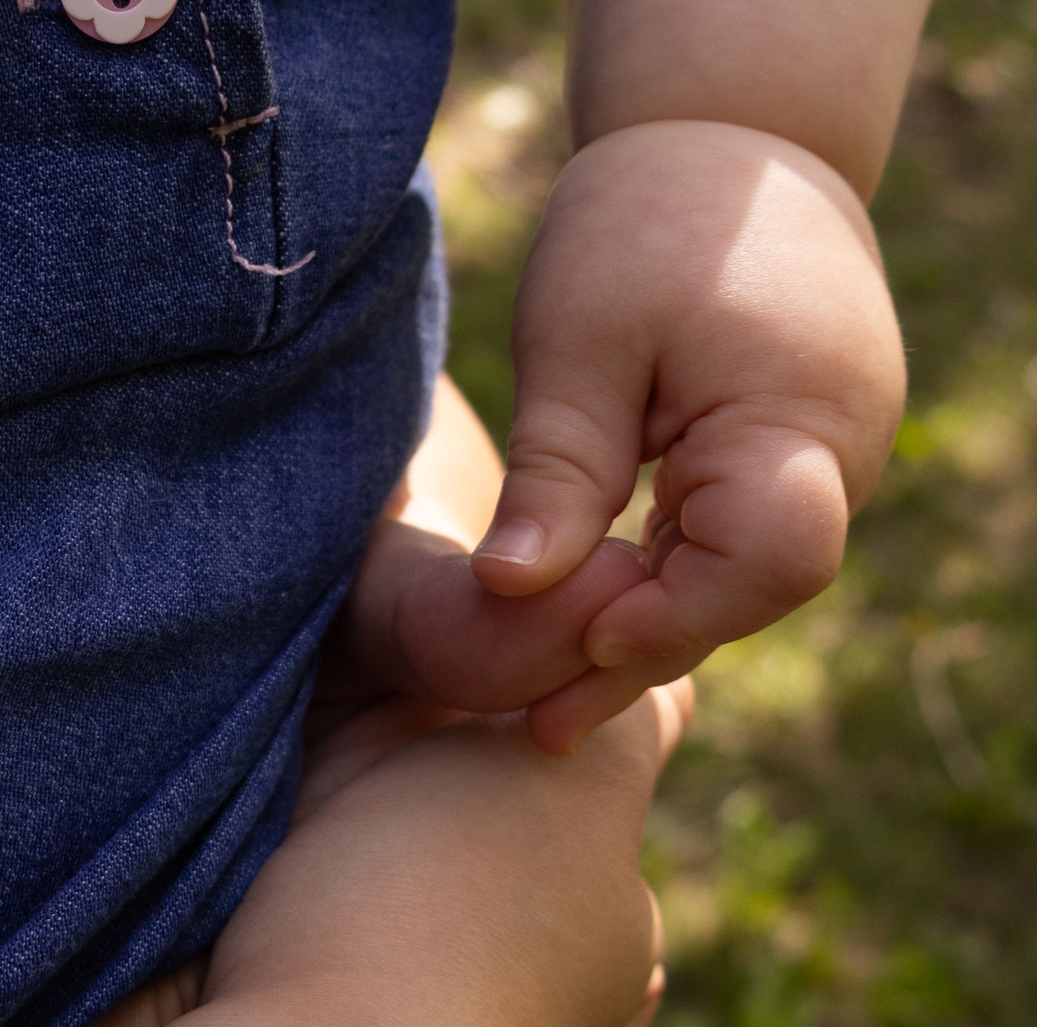 Close-up of a small baby hand gently grasping an adult finger against denim clothing.