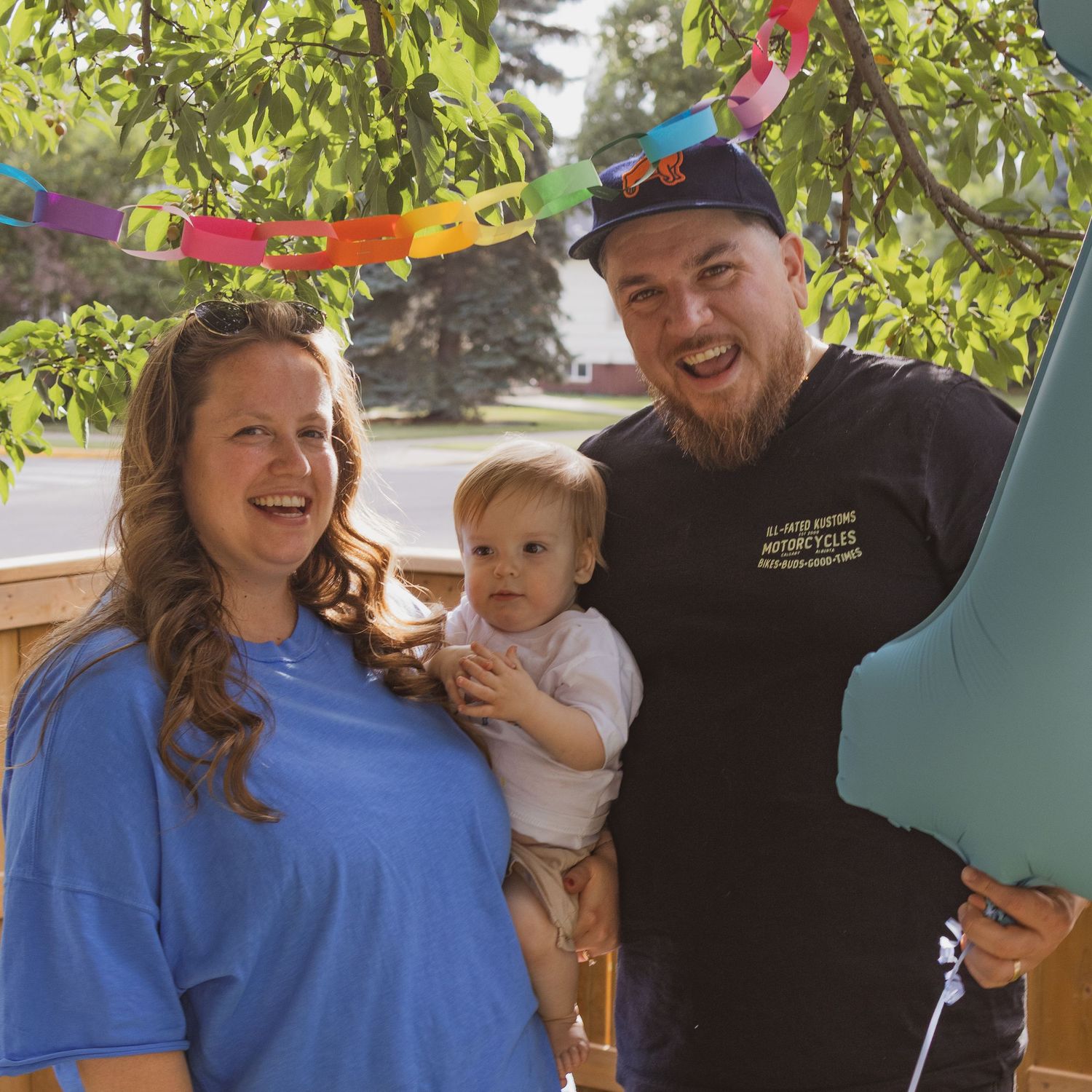 A smiling family of three with a baby poses outdoors under a tree with colorful paper chain decorations.