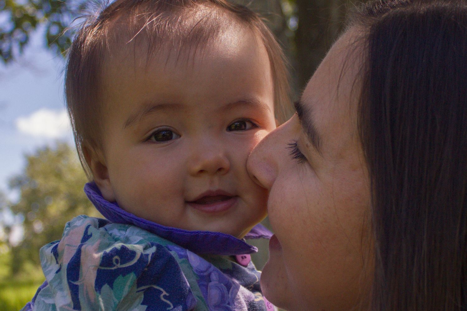 Mother kissing smiling baby in colorful outfit outdoors with trees in background.