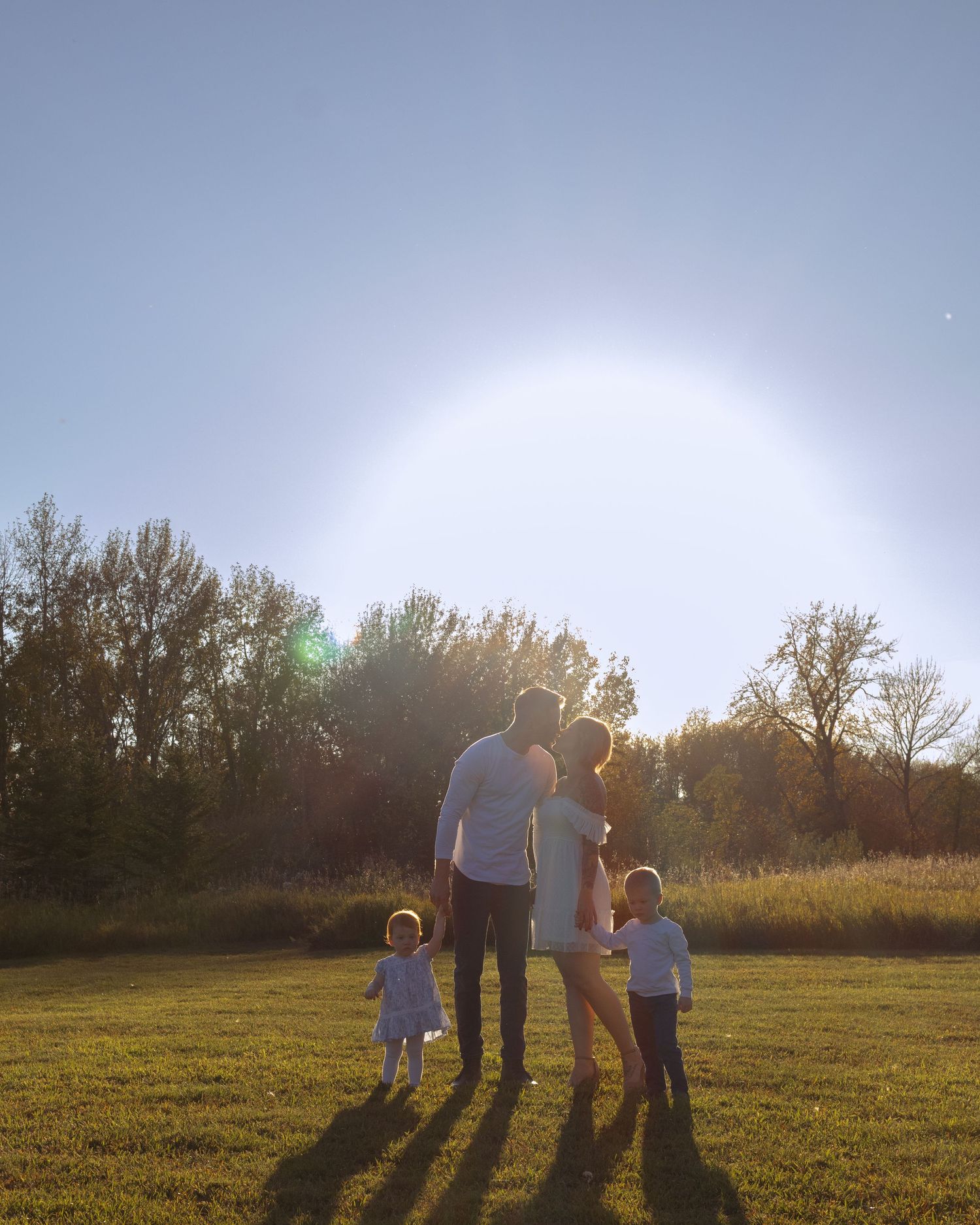 Family of four standing in a sunlit field at golden hour, with trees in the background and long shadows on the grass.