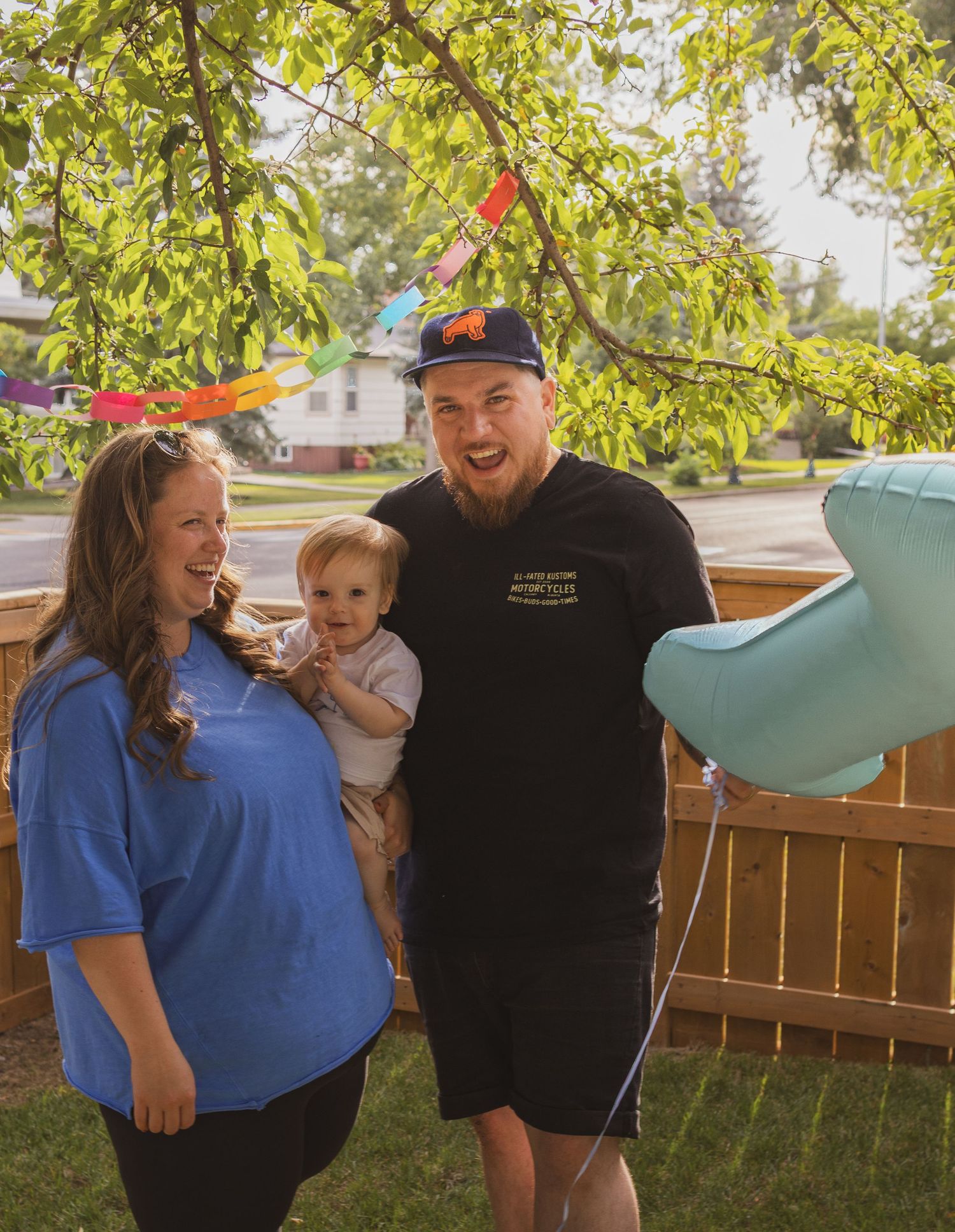 A family laughs outdoors holding a blue balloon under a tree with colourful paper chain garlands.