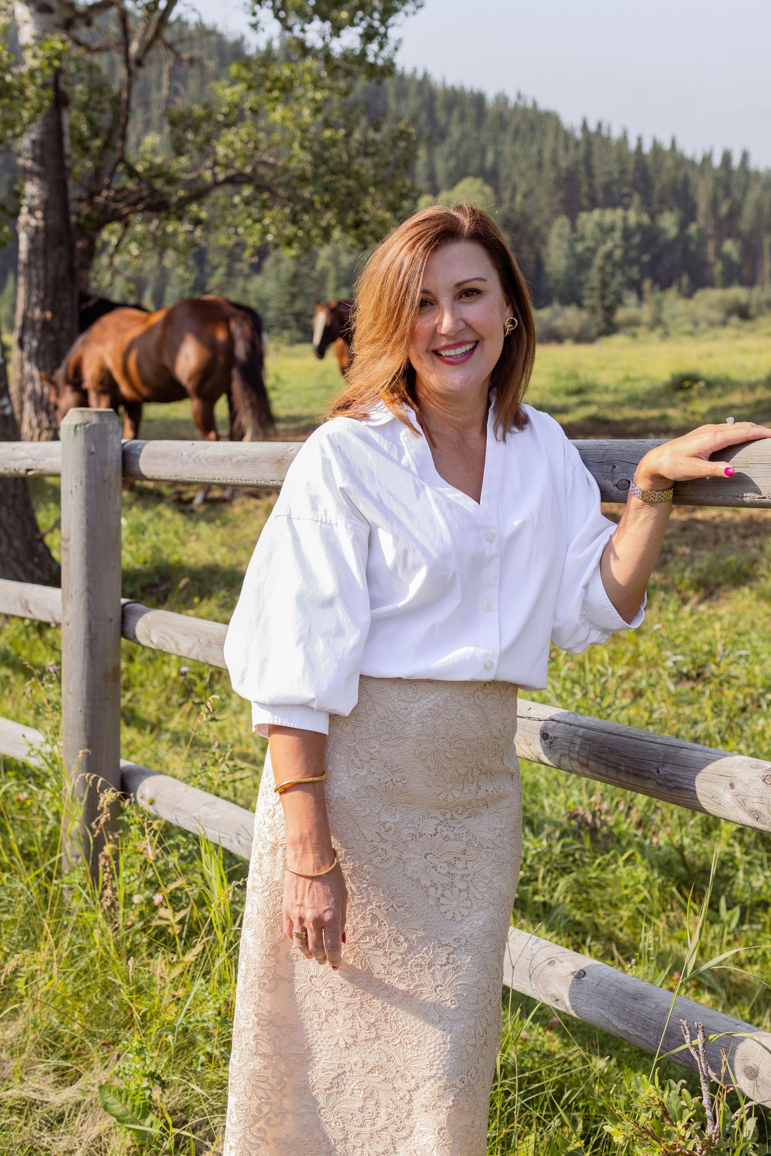 A smiling redhead woman in a white blouse and lace skirt poses by a wooden fence with horses grazing.