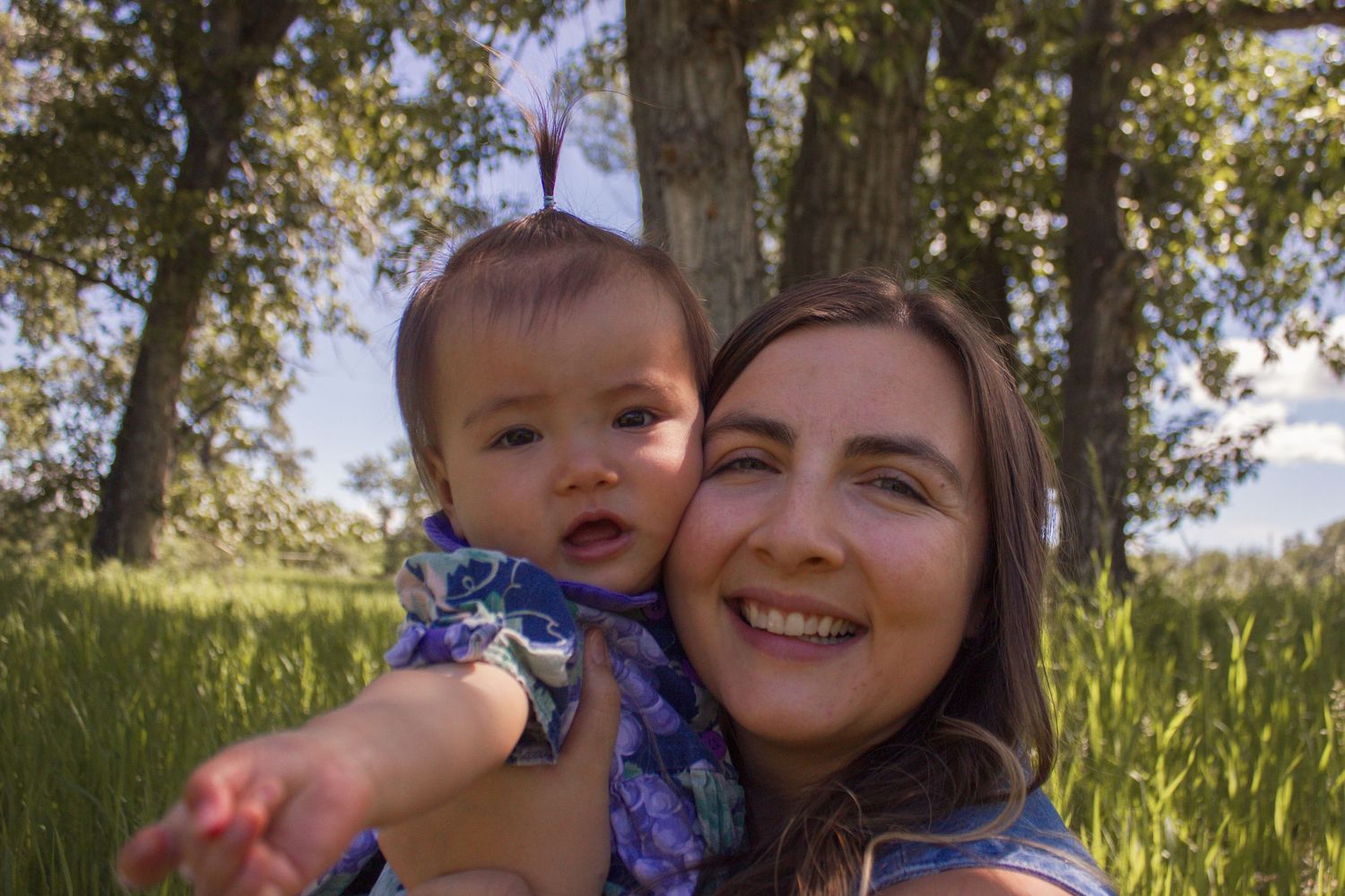 A happy mother holds her baby girl up outdoors surrounded by tall green trees and grass.