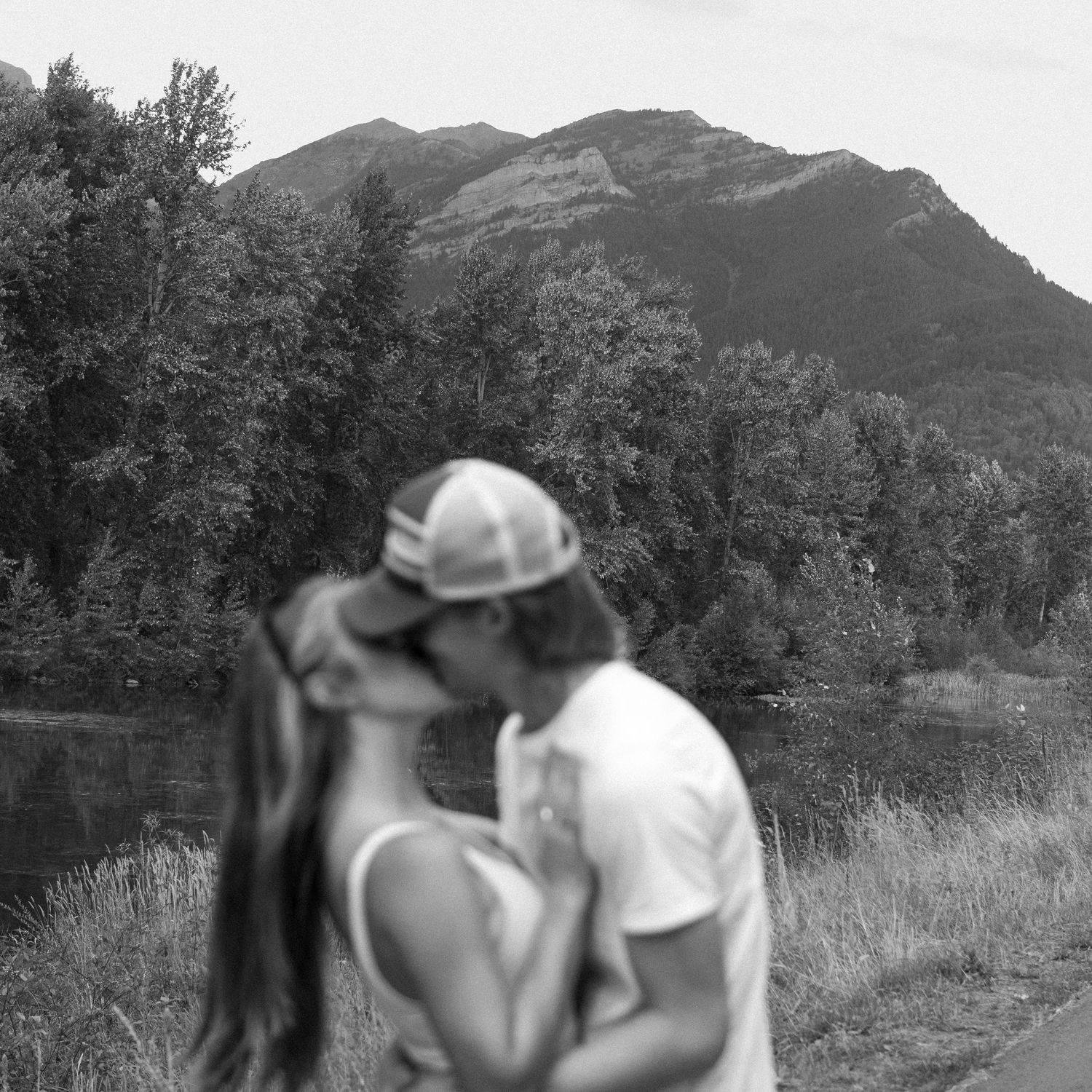 Black and white photo of a couple kissing near a mountain lake surrounded by trees and tall peaks.