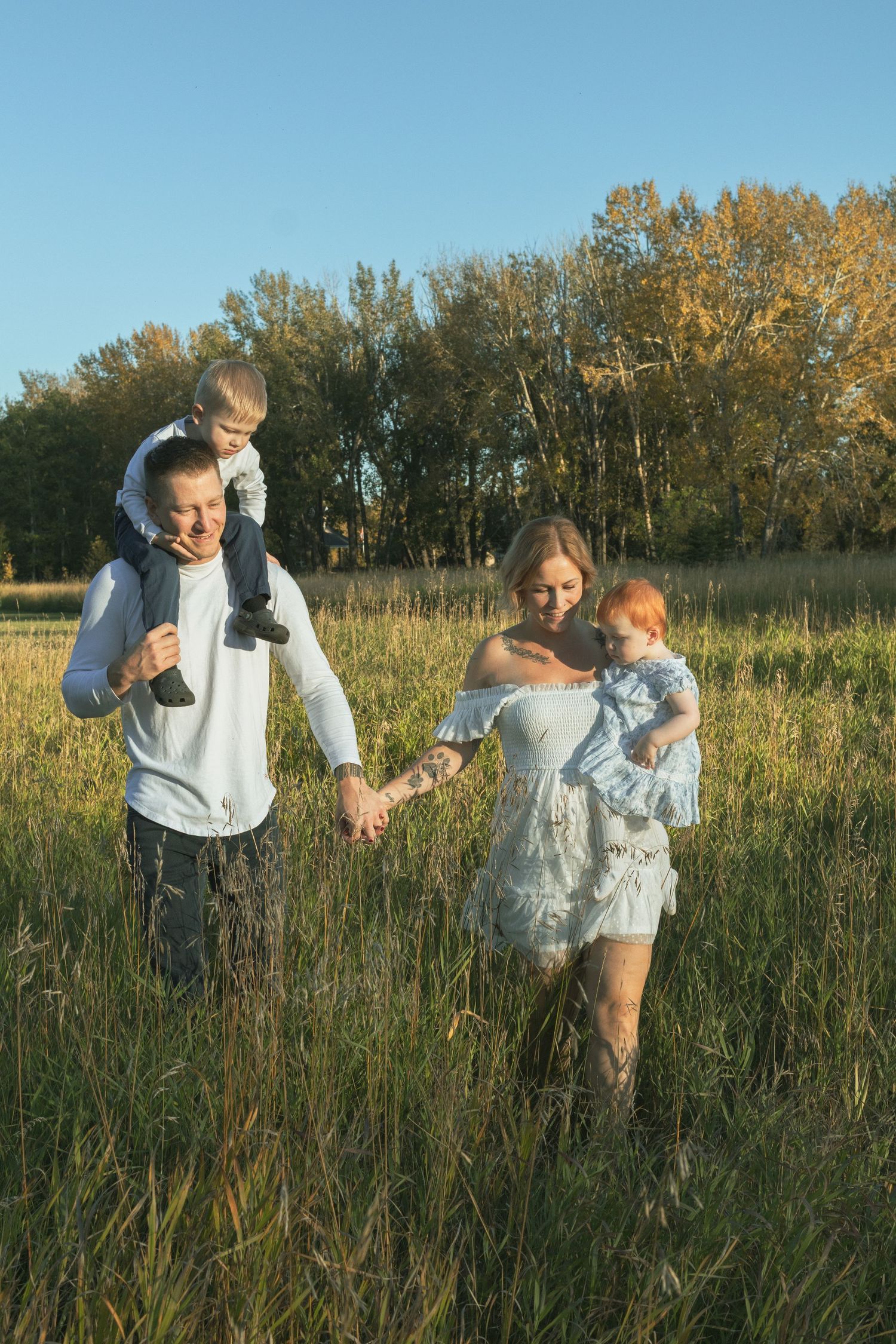 Happy family walking in a sunny field, father carrying son on shoulders, mother holding baby, autumn trees background.