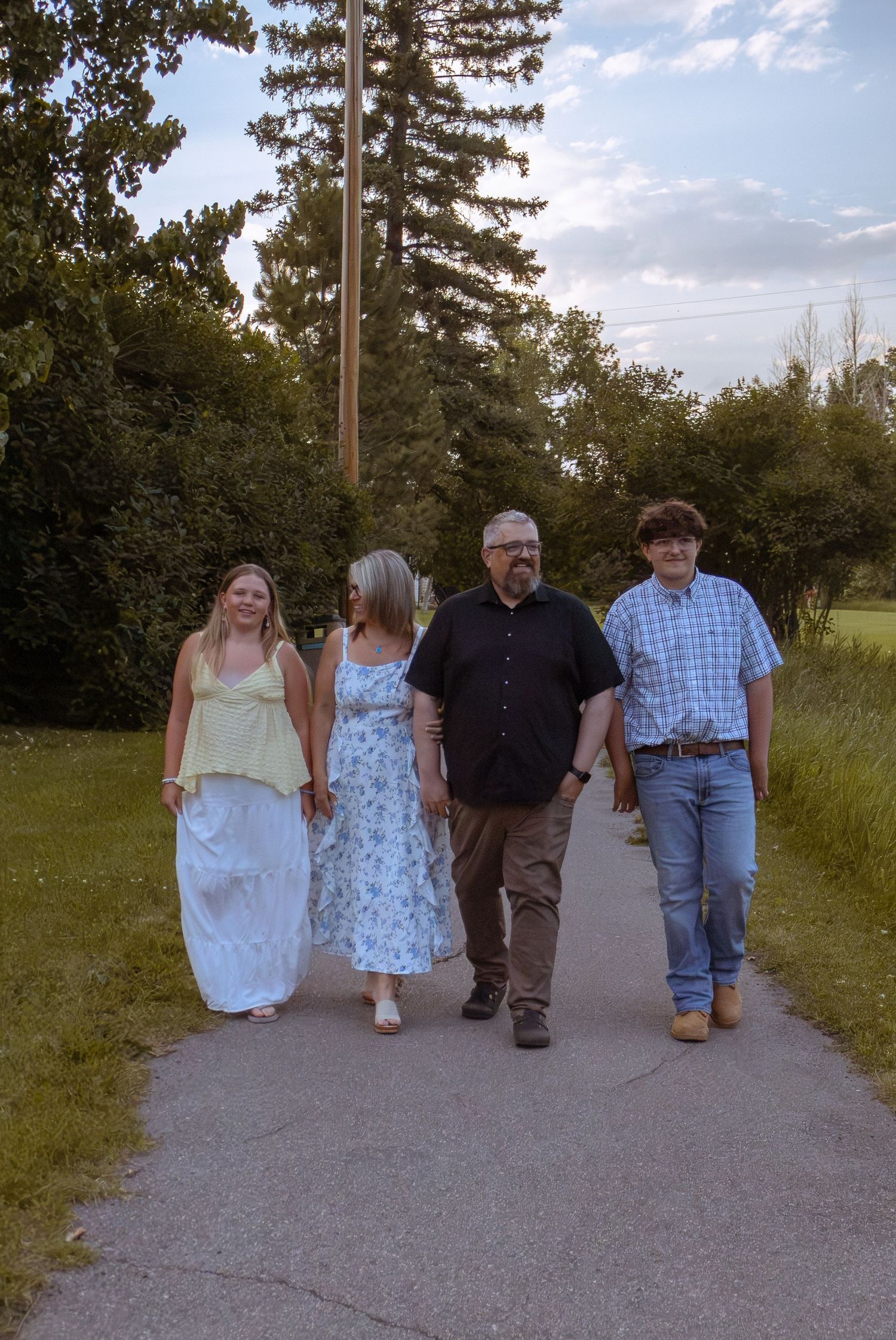 Family of four walking together down a sunny park path surrounded by green trees and grass.