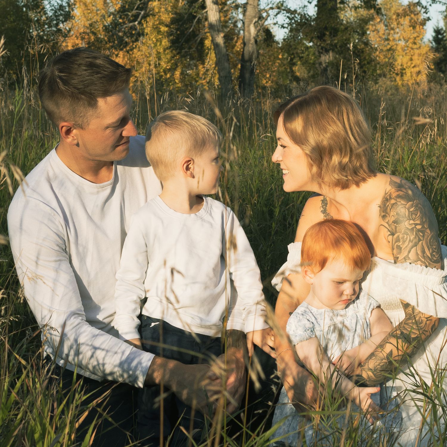 Family of four sitting in golden meadow grass, parents in white with tattooed arms, two young children, warm sunset light.