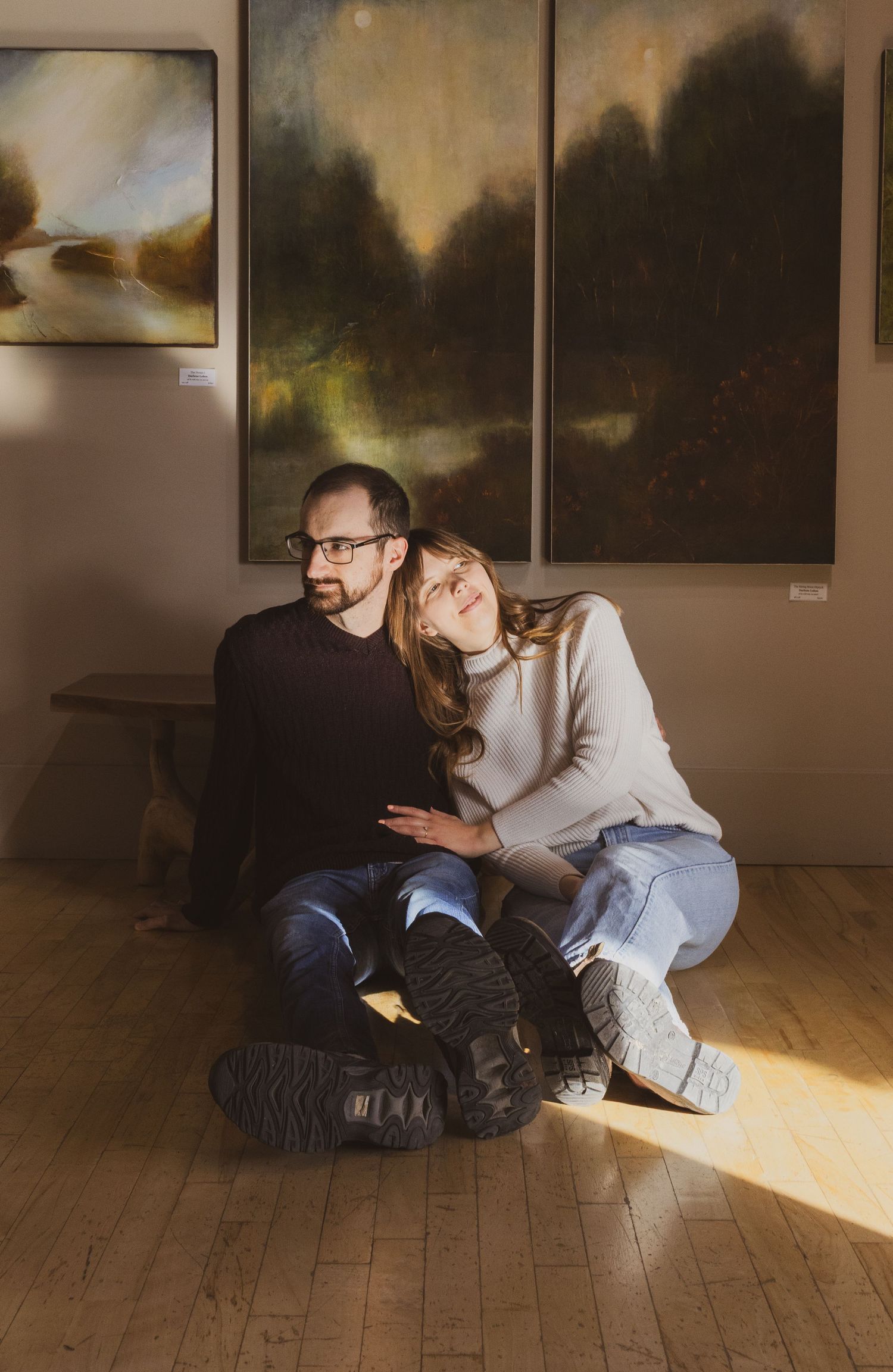 Couple sitting on wooden floor in art gallery, surrounded by dark landscape paintings, sharing a tender moment in warm sunlight.