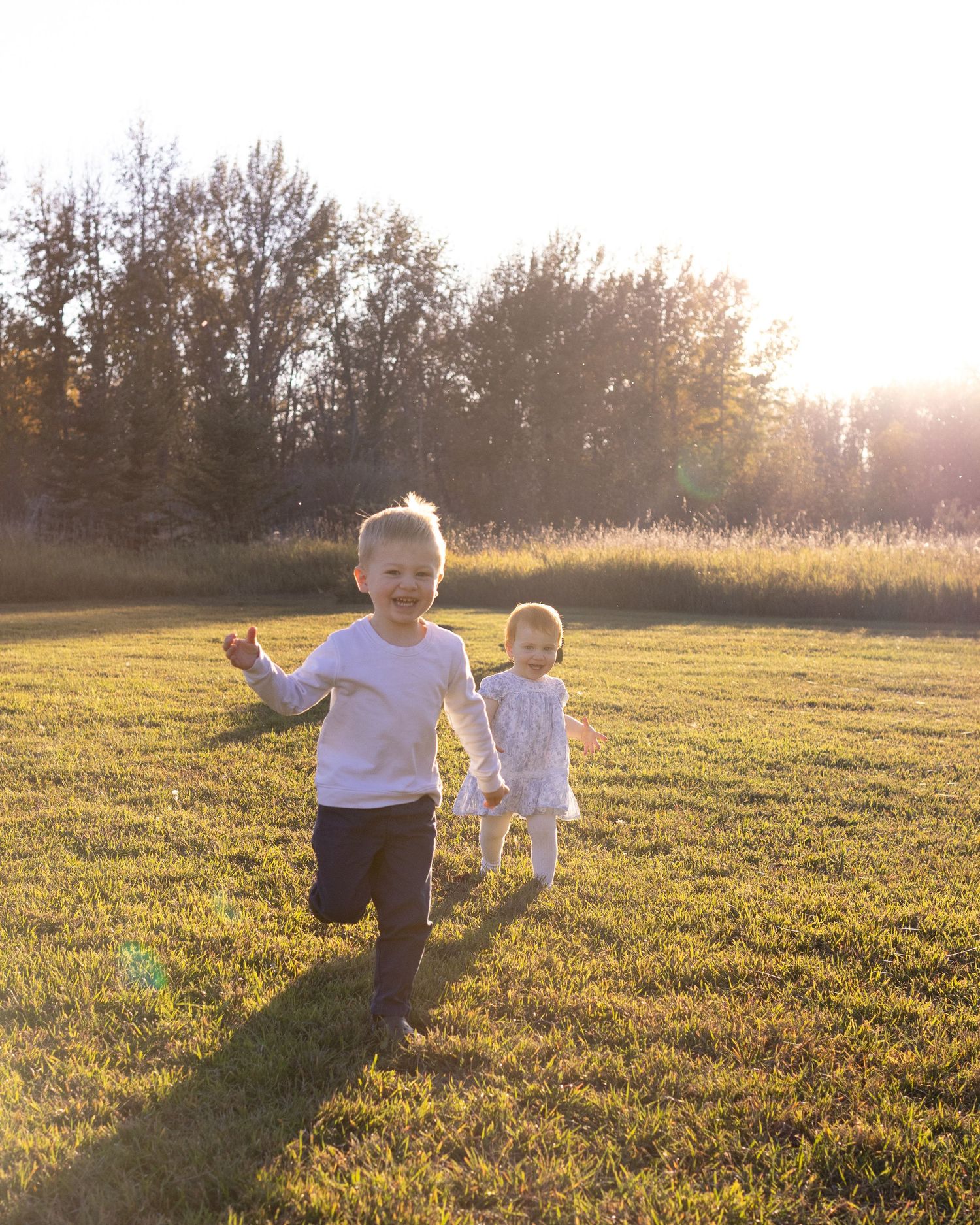 Two young children running and laughing in a sunny green field with golden trees in the background.