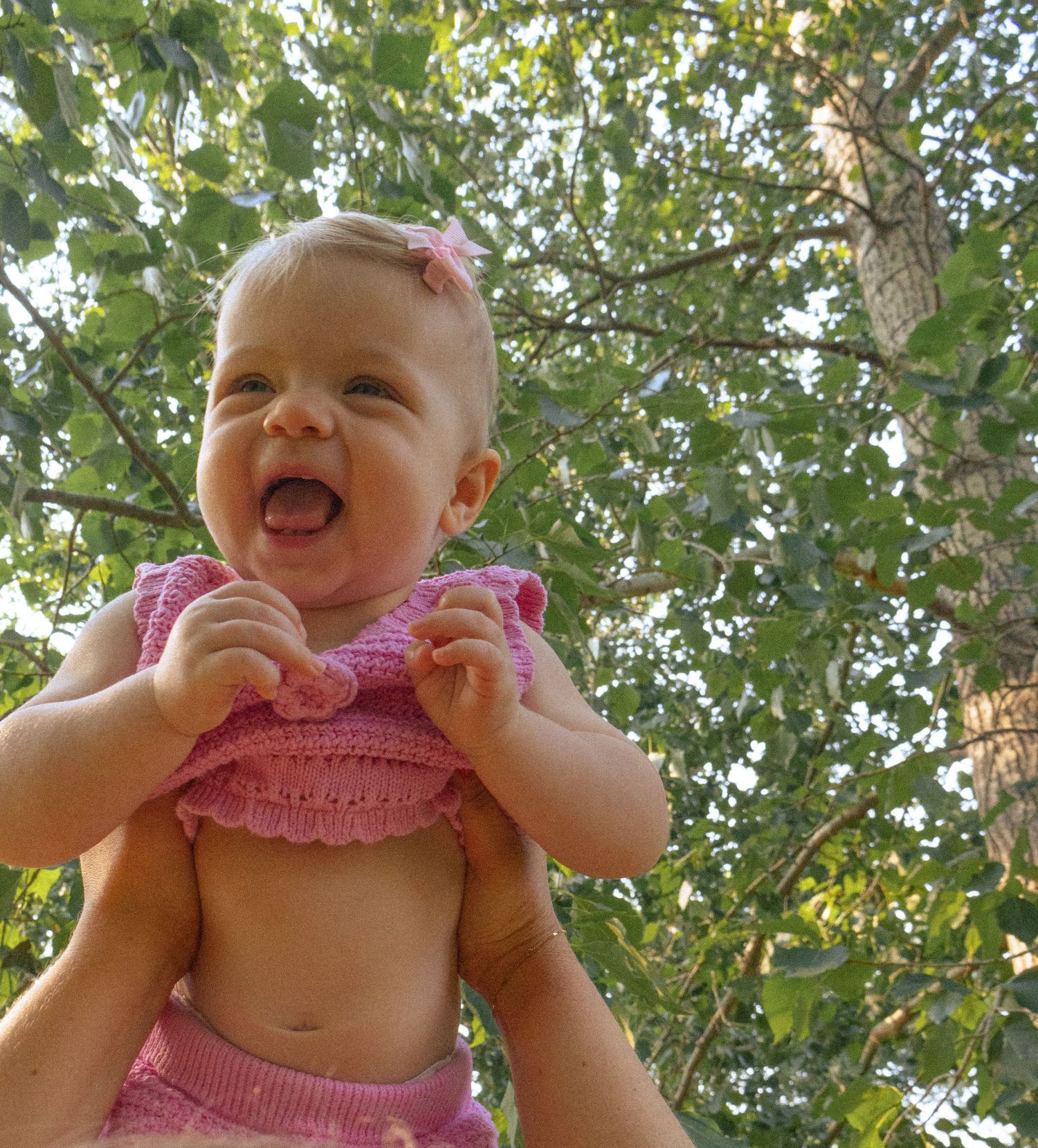 Happy laughing baby in pink knit outfit being lifted up outdoors with green trees in background.