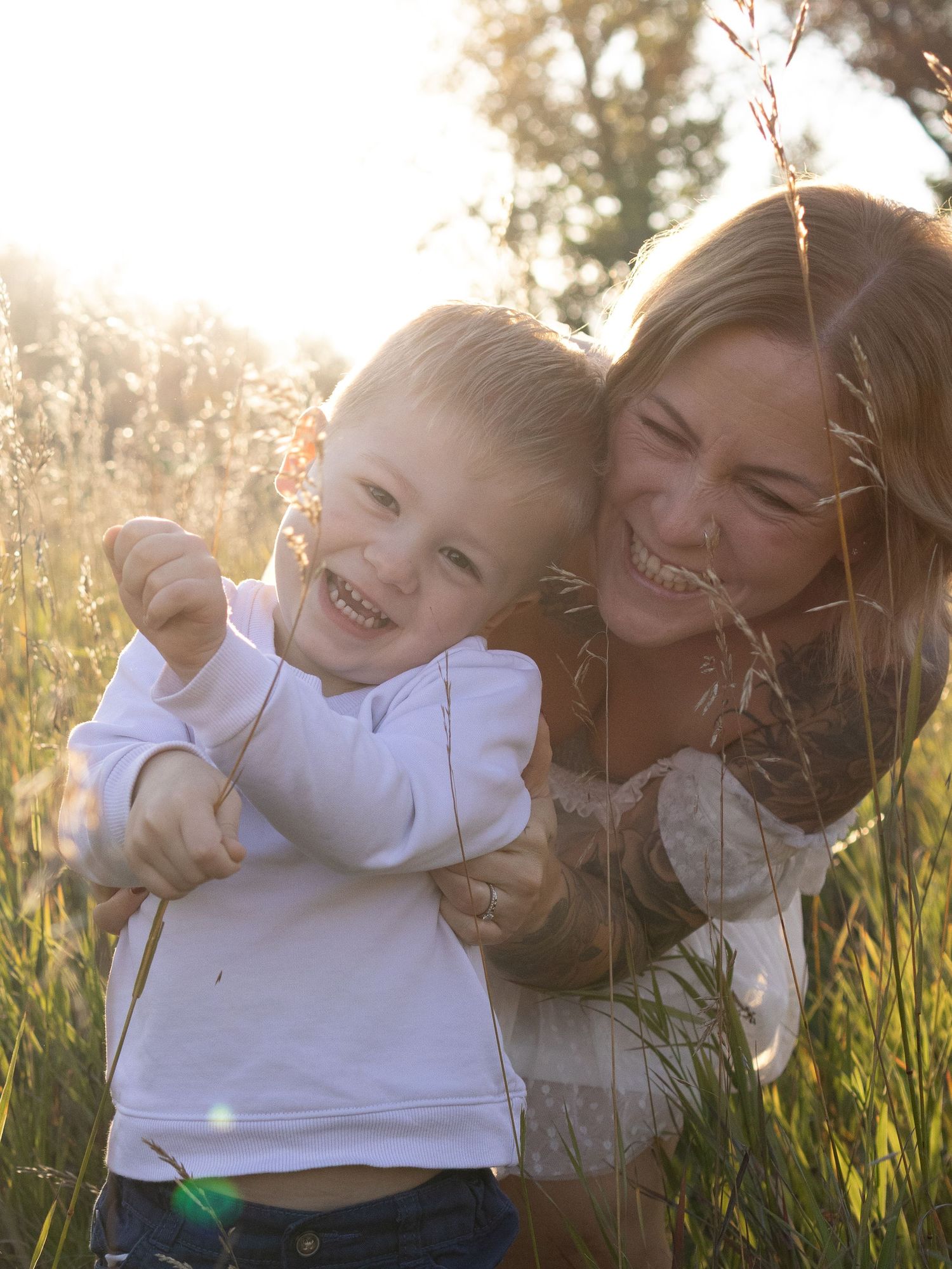 A joyful mother hugs her laughing toddler boy in a sunlit meadow with golden backlight and tall grass.