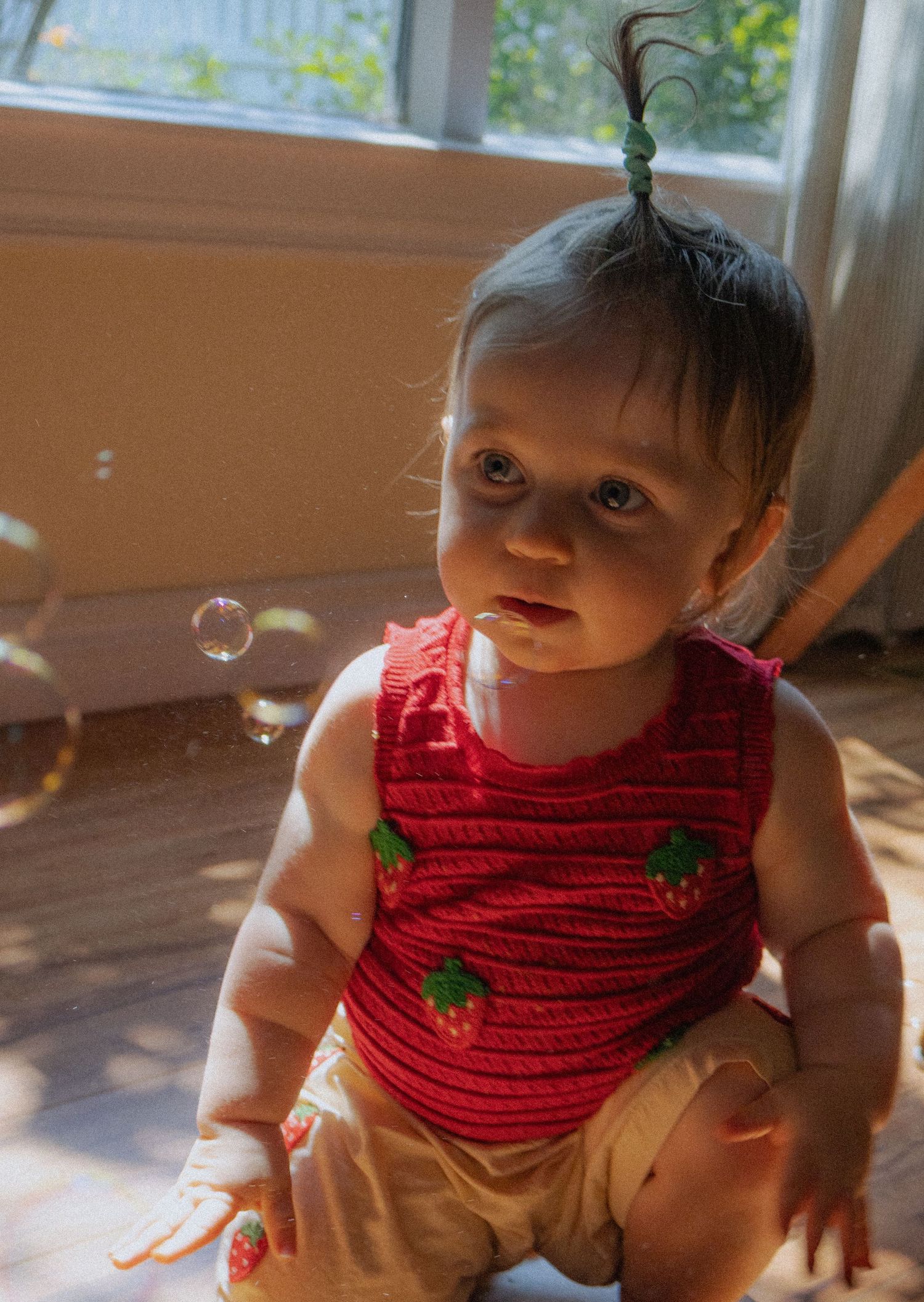 Curious baby in red strawberry knit top sitting on floor with soap bubbles floating nearby indoors.