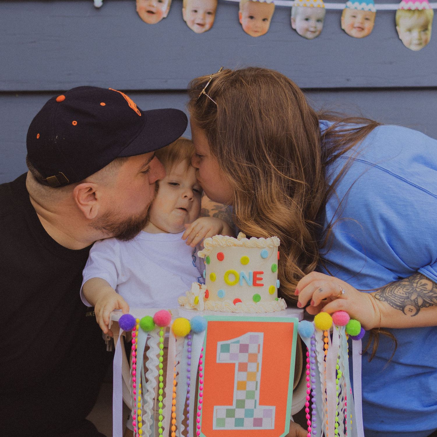 Two parents kiss their toddler on the cheeks while the child holds a colorful first birthday cake.