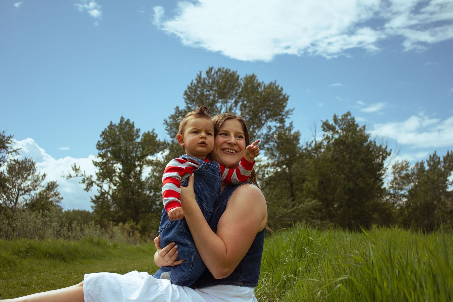 A mother holds her baby in a green field under a bright blue sky.