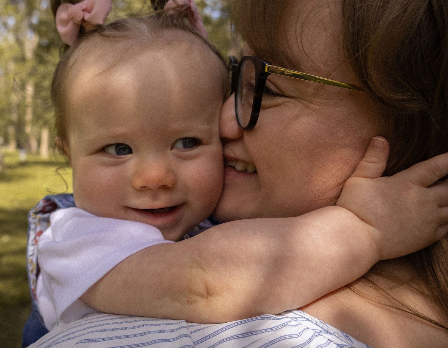 Happy baby smiling while being hugged and kissed by woman wearing glasses in sunny park.