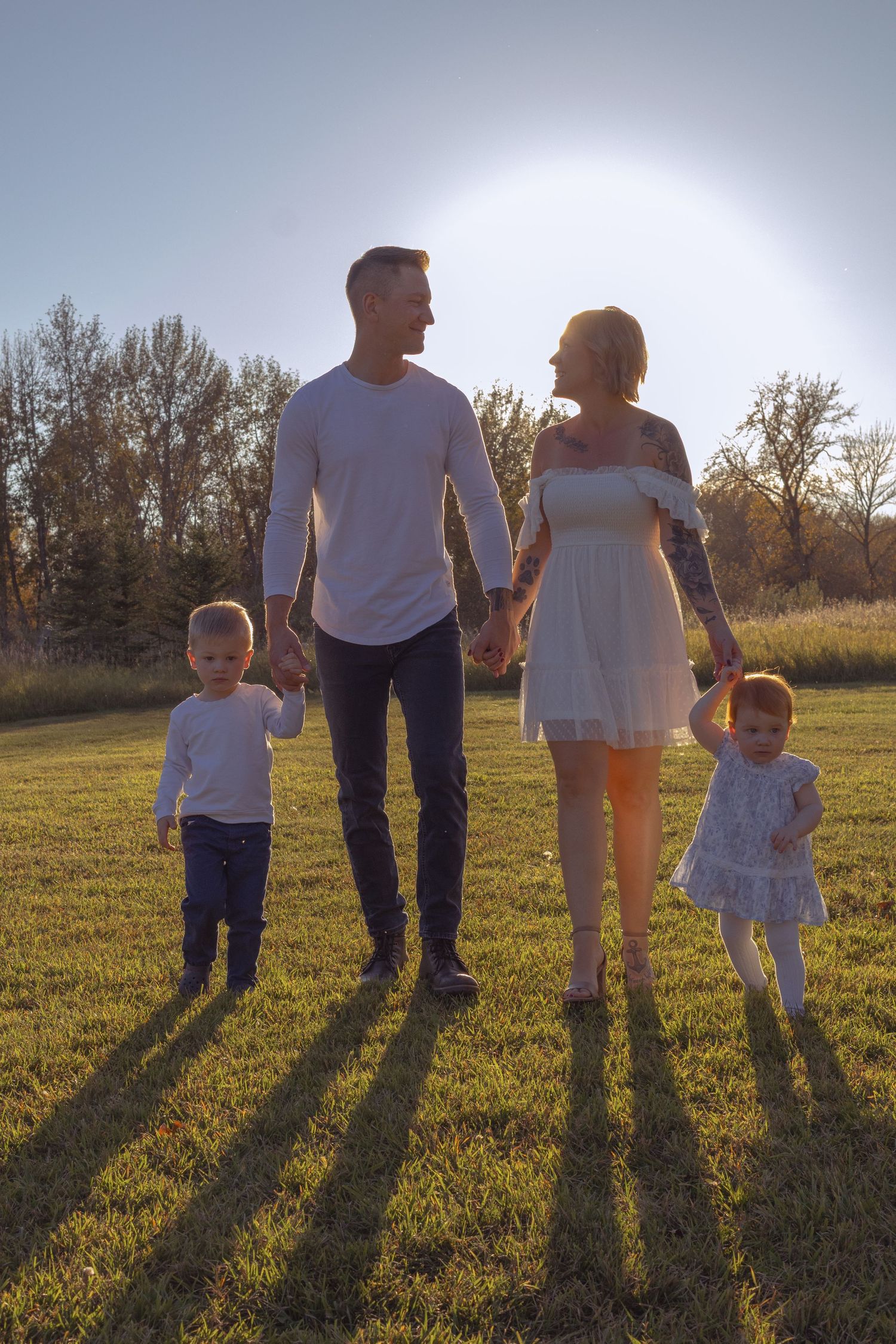 Family of four holding hands in a sunny park, parents and two young children smiling outdoors at golden hour.