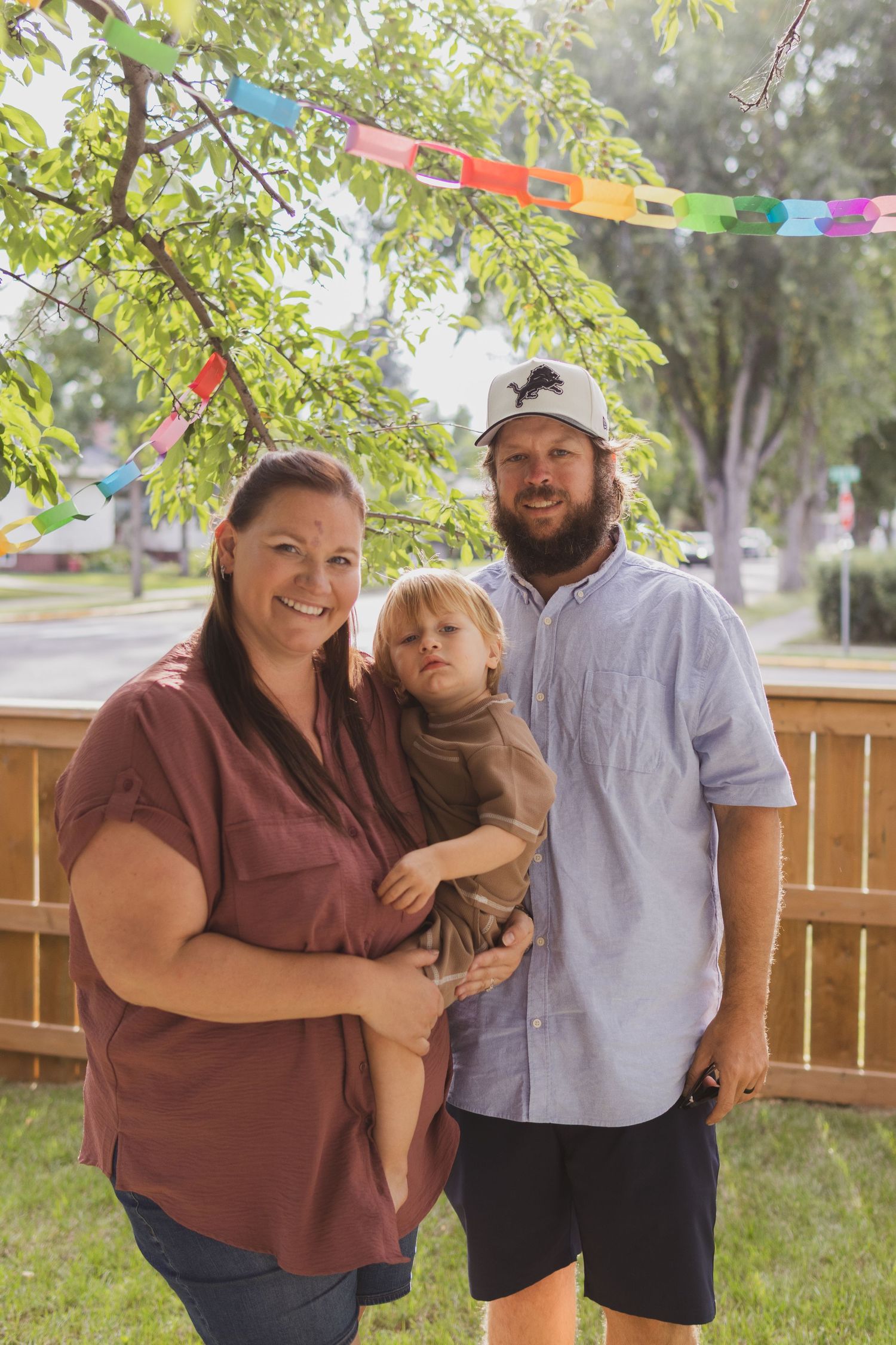 A smiling family of three poses outdoors with colorful bunting, woman holding toddler beside bearded man in cap.