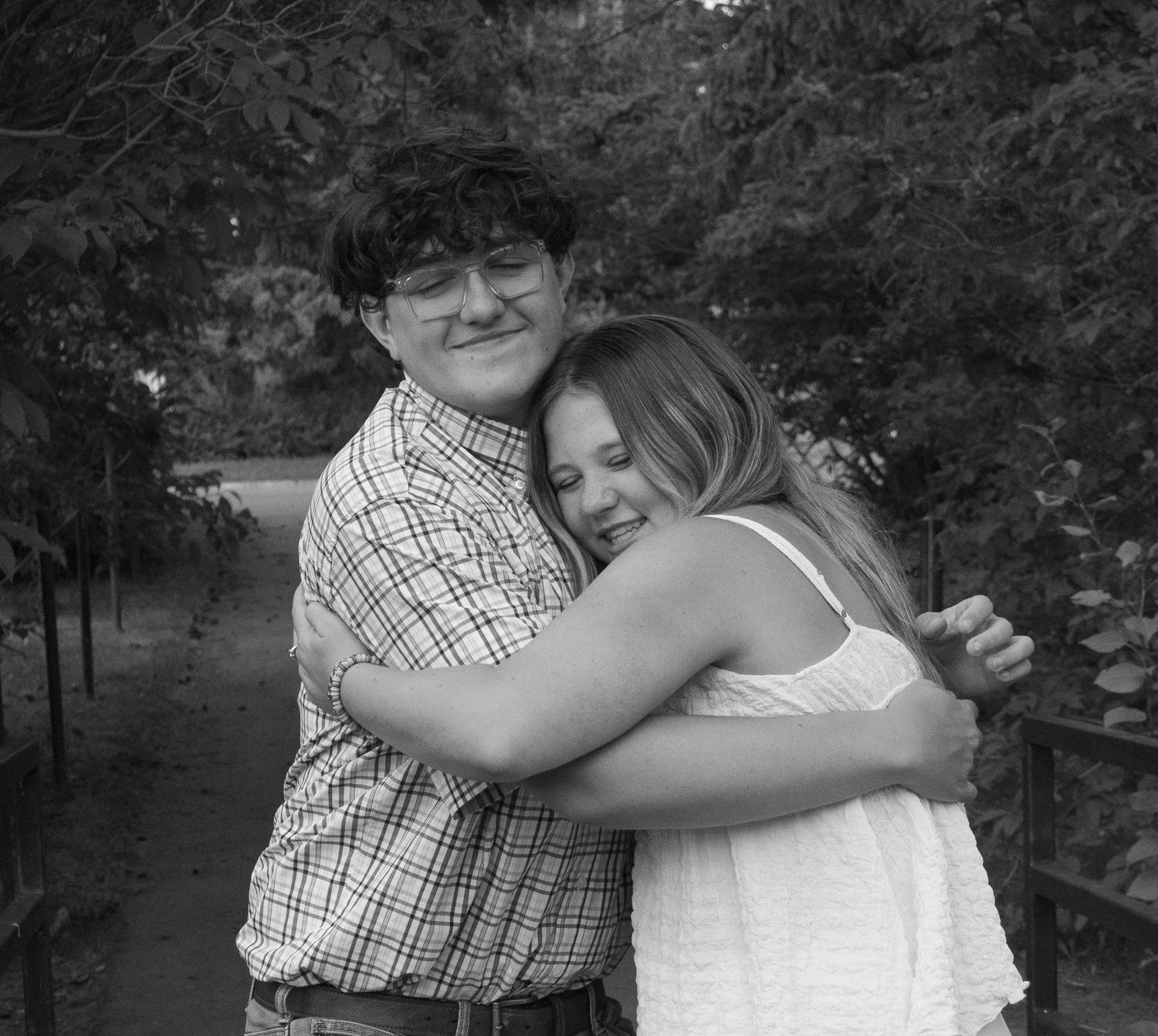 Black and white photo of a teenage boy and girl sharing a warm hug on an outdoor bridge.