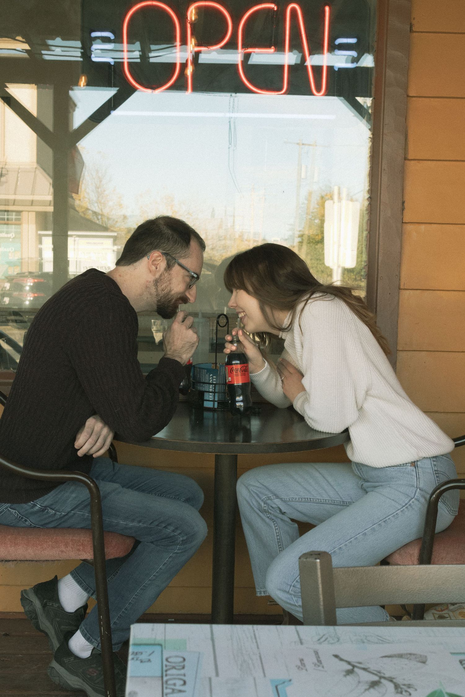 Couple sitting across from each other at a small cafe table near an OPEN neon sign, sharing an intimate moment.