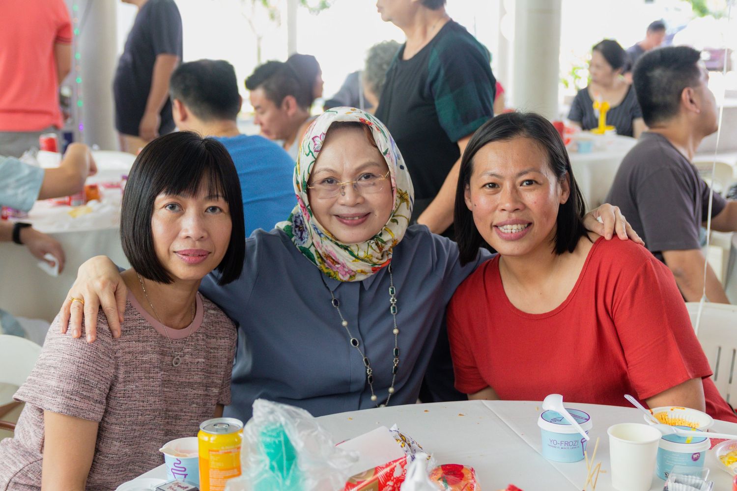 Three smiling women pose together at a table during a casual indoor gathering in a busy dining area.
