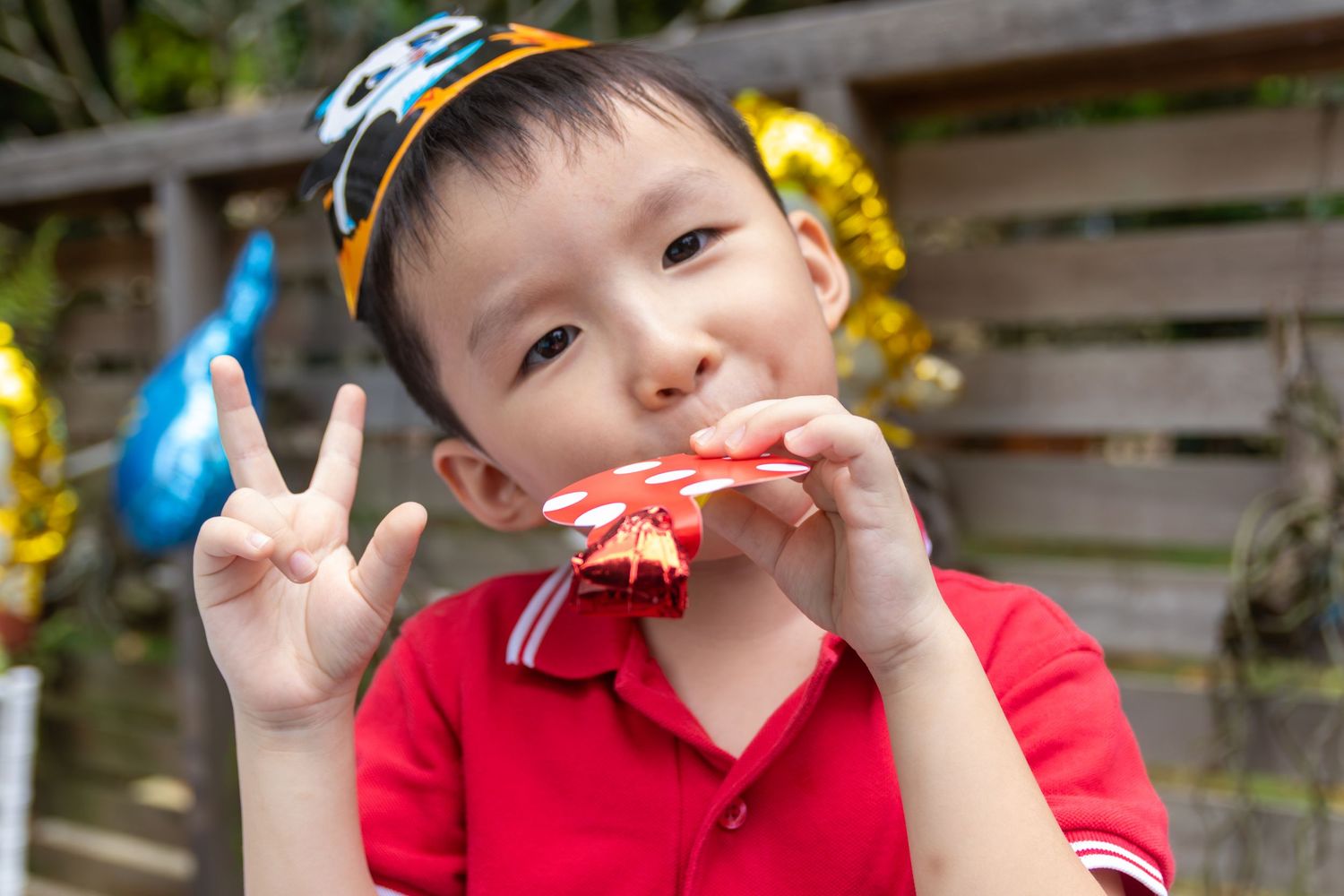 Young boy in party hat making peace sign while holding a red toy at an outdoor birthday celebration.