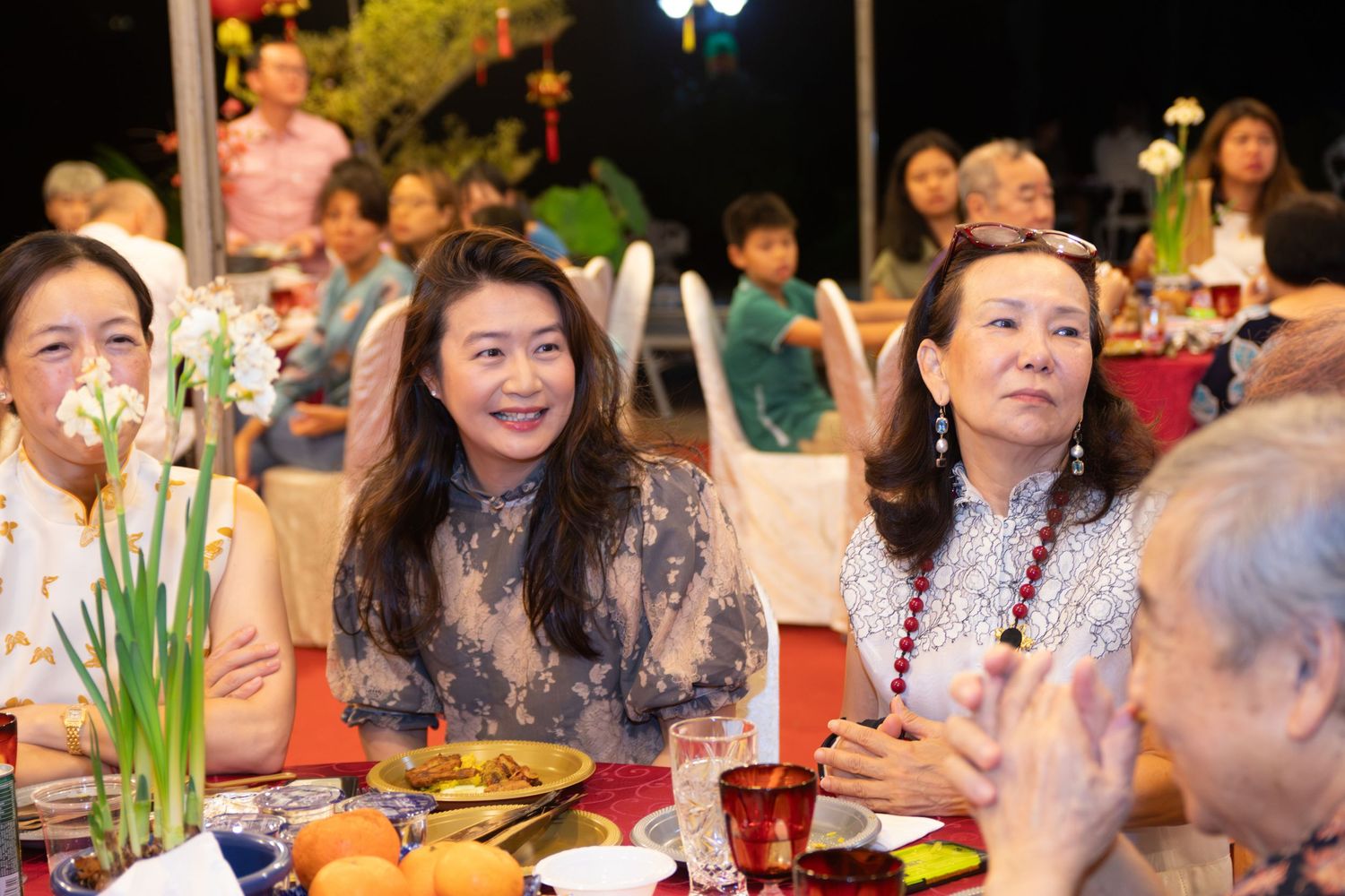 Women smiling at a festive Asian dinner gathering with flowers, fruit, and traditional decorations on the table.