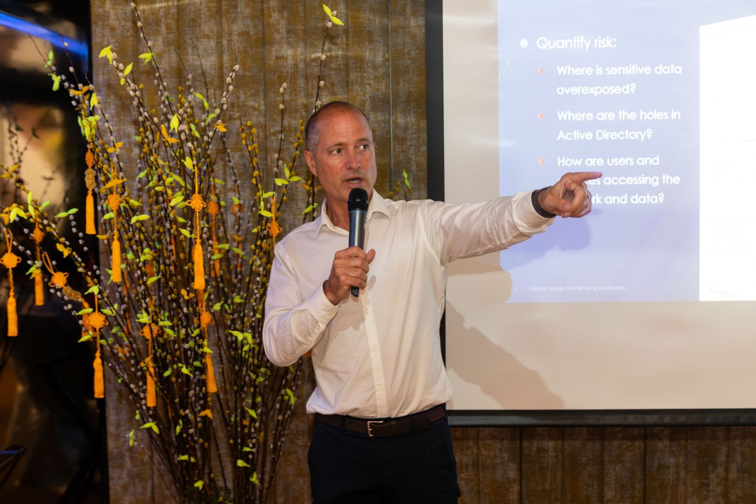 A man in a white shirt speaks into a microphone and points at a presentation screen at a corporate event.