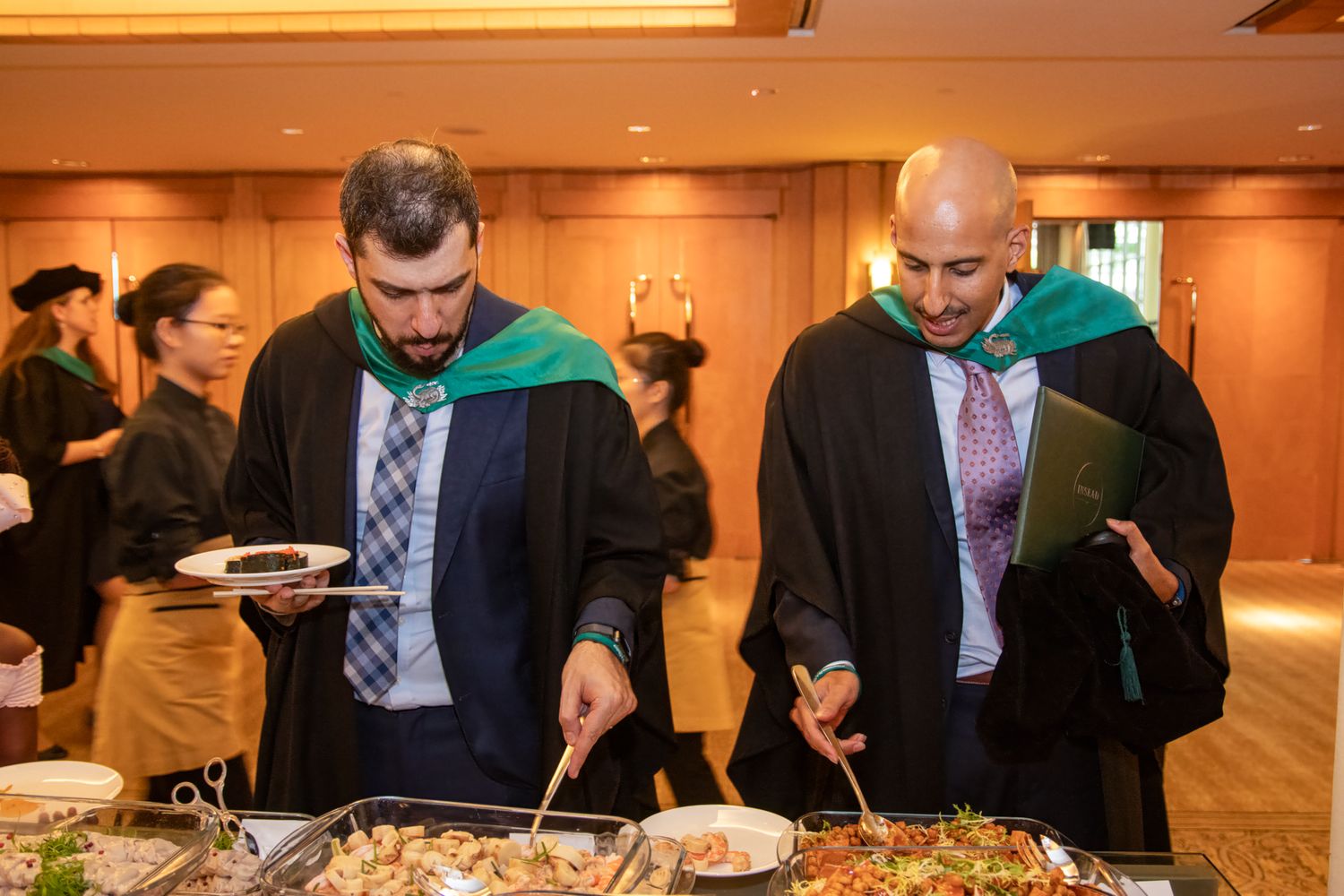 Two graduates in academic robes with green hoods serve themselves at a buffet during a graduation reception.