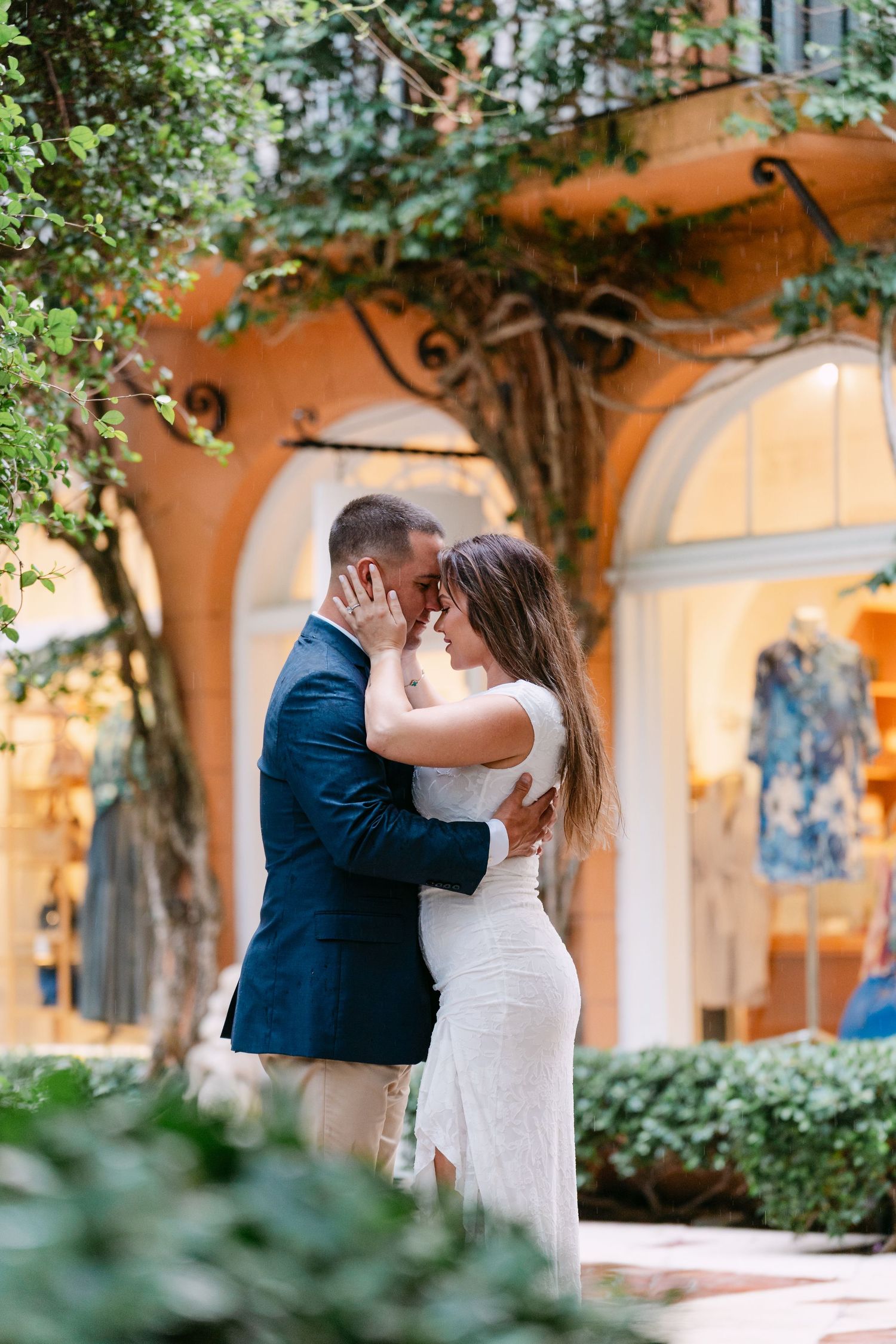 Couple embracing intimately outdoors surrounded by lush greenery near a warmly lit boutique storefront.