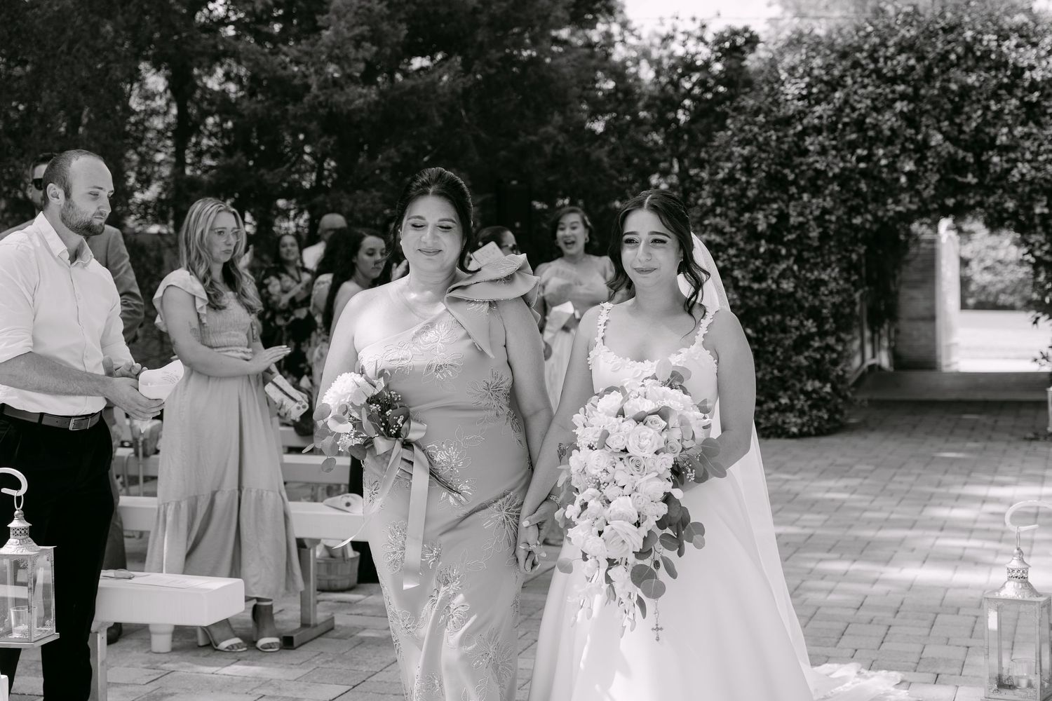 Bride in white gown walks with bridesmaid in elegant outdoor wedding ceremony, black and white photo.
