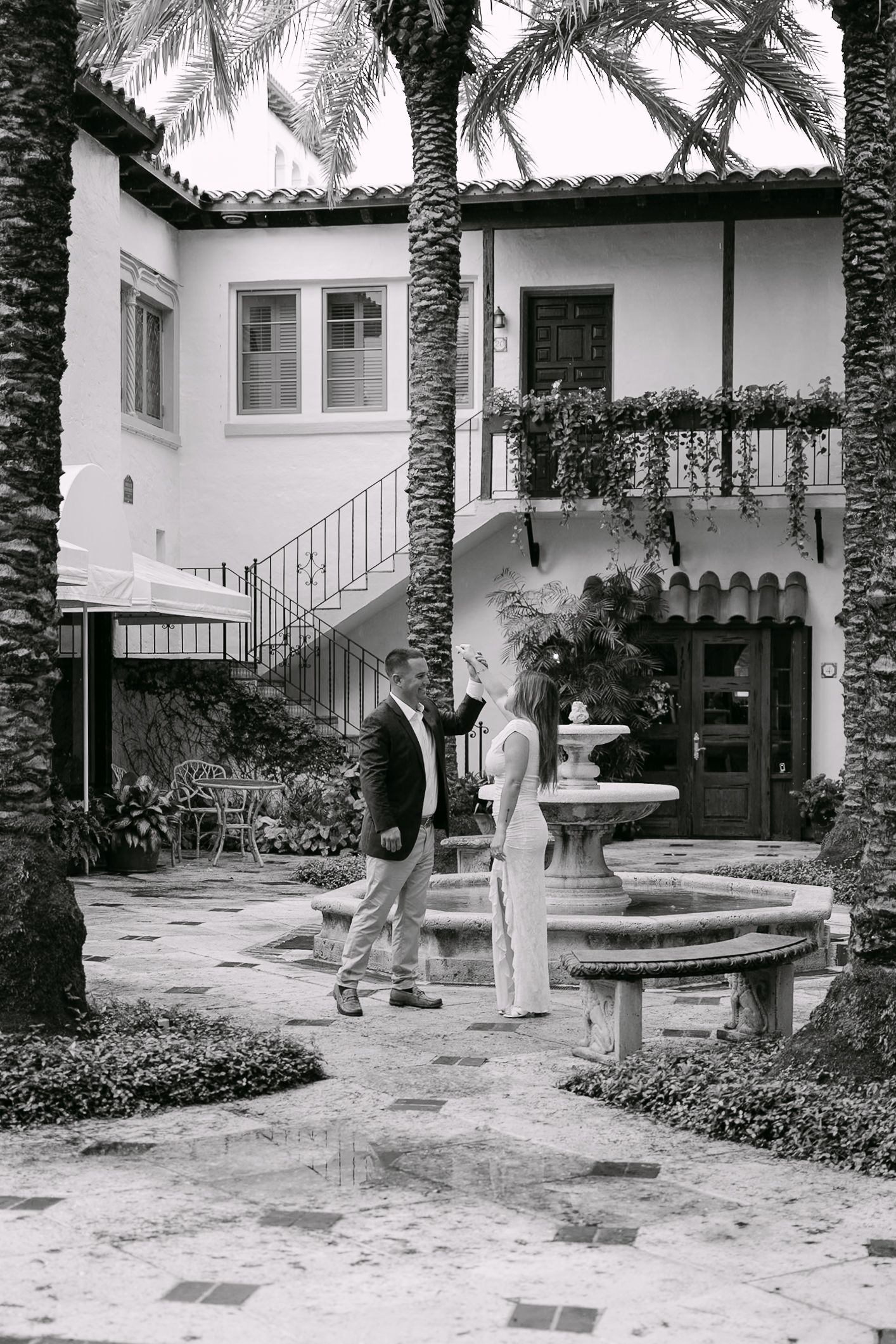 Black and white photo of a couple conversing by an ornate fountain in an elegant courtyard with palm trees.