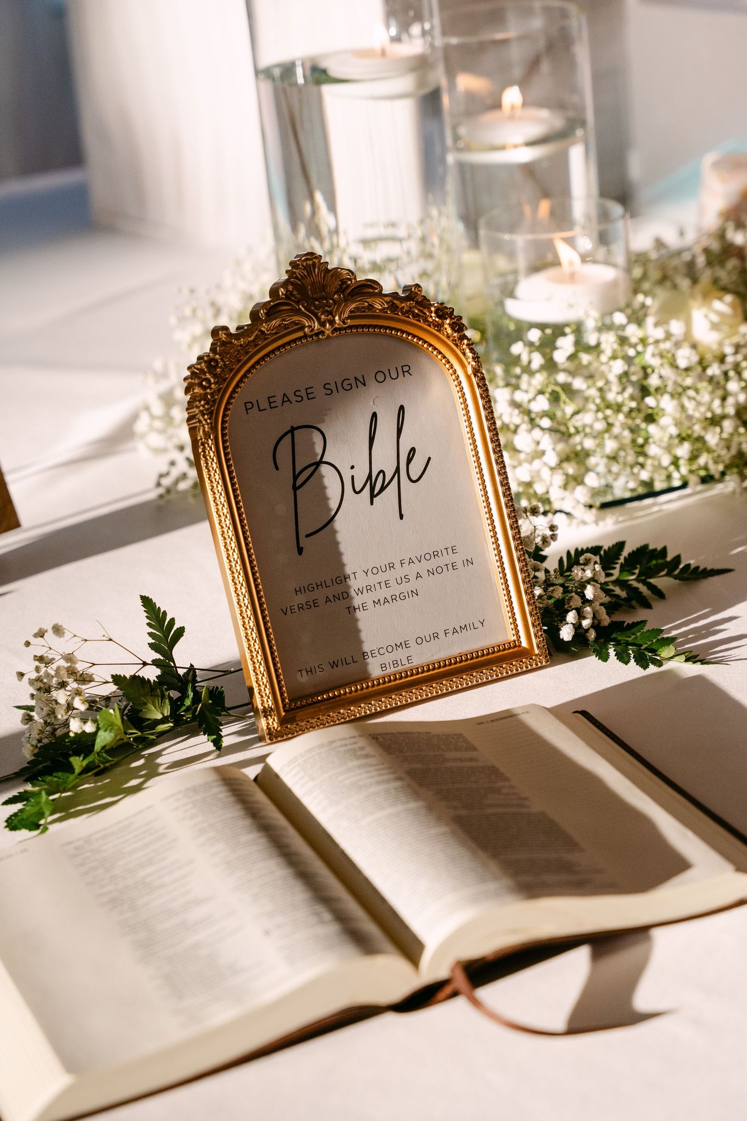 Elegant wedding Bible signing station with gold framed sign, candles, and white floral greenery decor.
