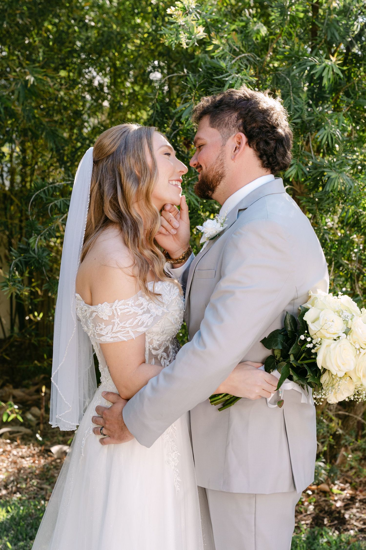 Bride and groom embrace and laugh together outdoors surrounded by green trees.