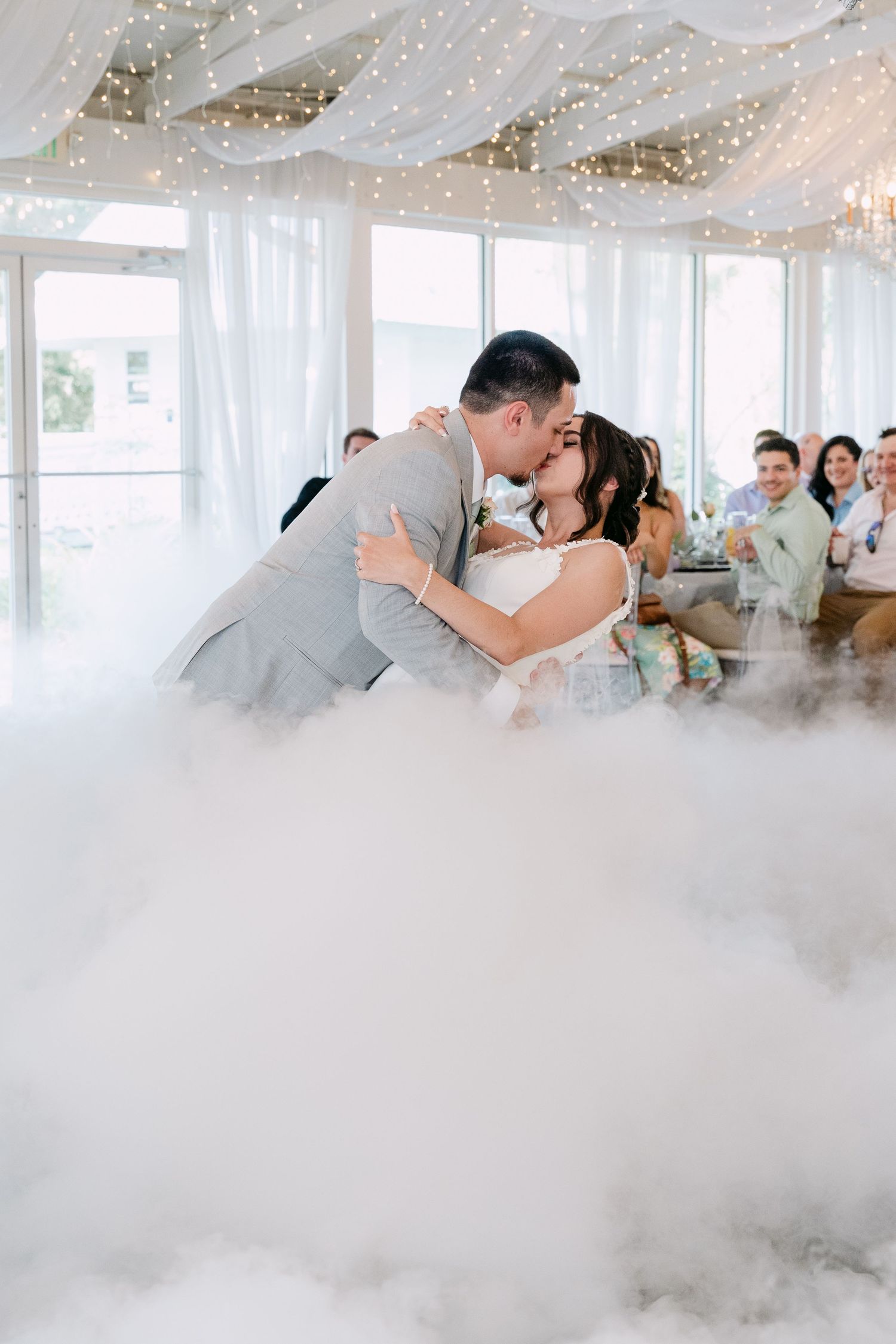 Bride and groom share a romantic first dance kiss surrounded by dramatic fog machine clouds at their elegant wedding reception.