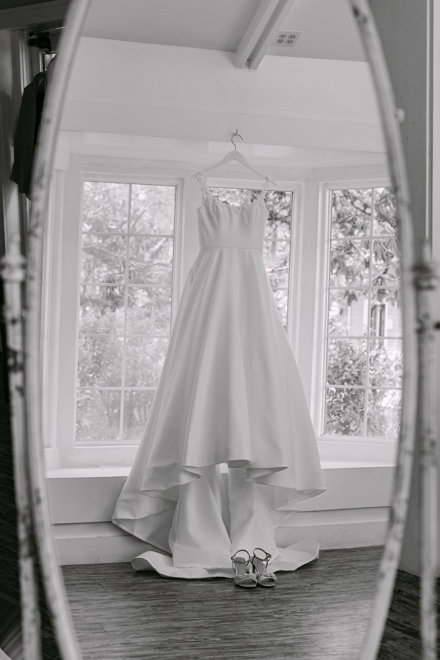 Black and white photo of an elegant strapless ball gown wedding dress hanging near bright windows with heels below.