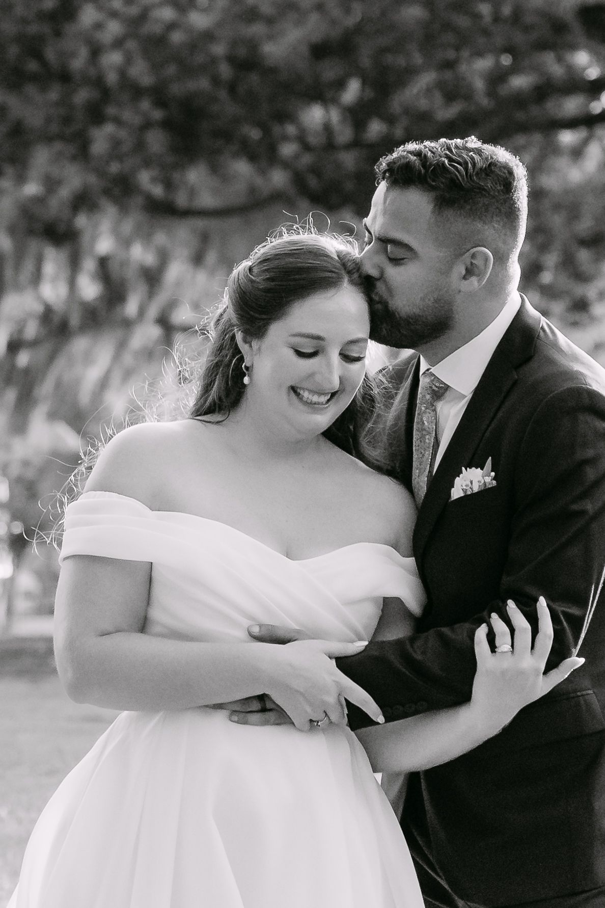 Black and white portrait of groom kissing smiling bride's cheek in an outdoor wedding setting.