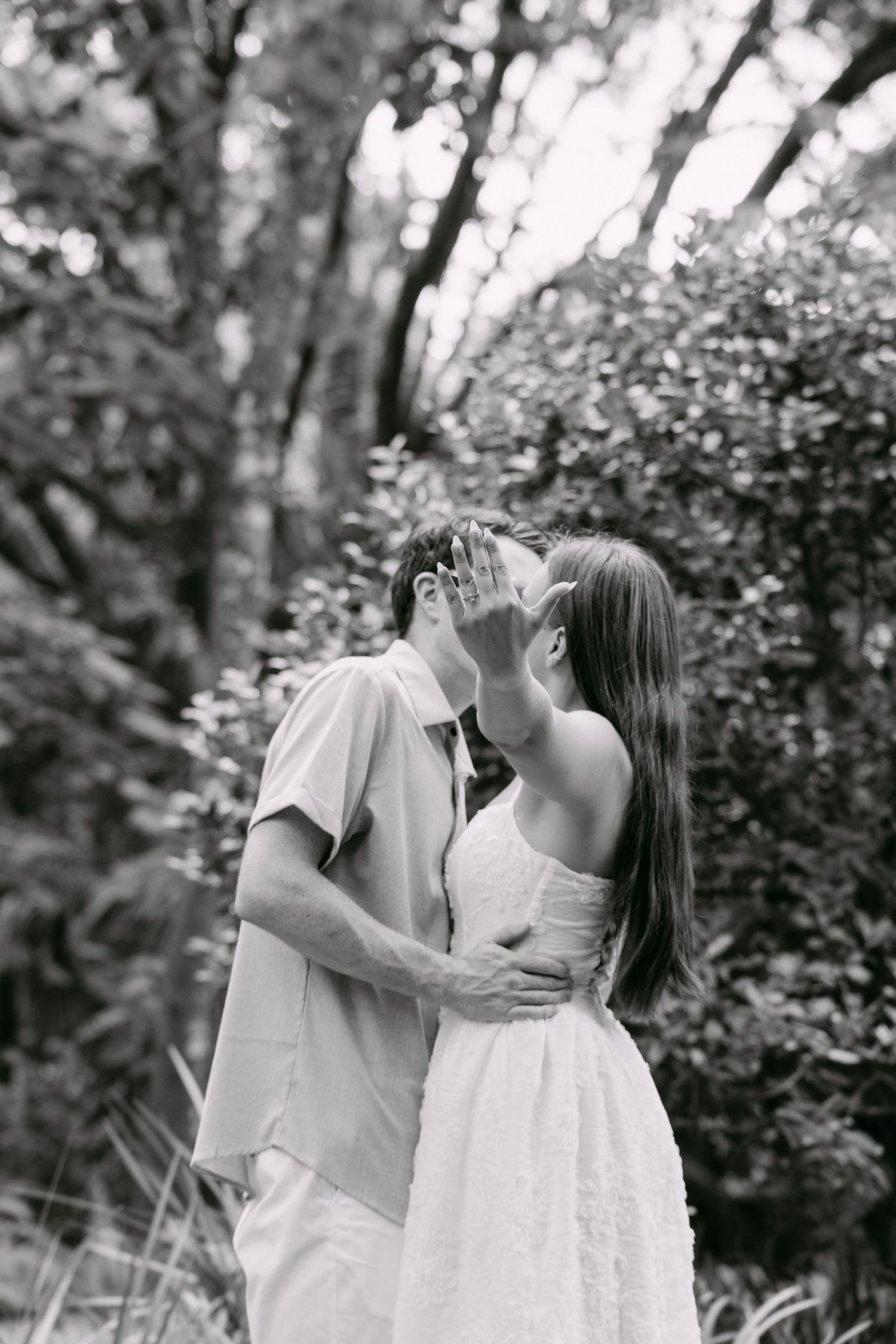 Black and white photo of a couple sharing a kiss outdoors surrounded by lush green trees and foliage.
