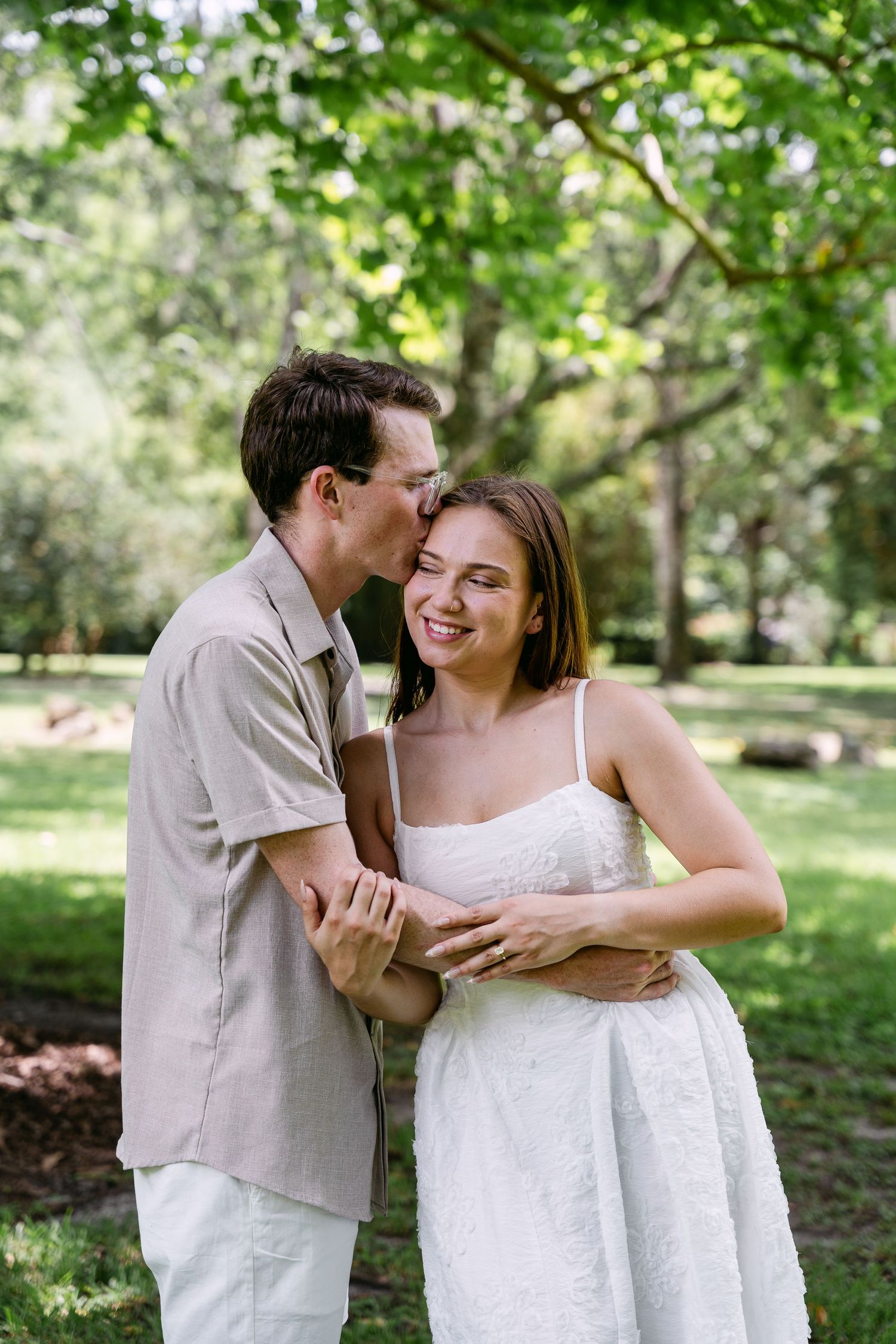 A couple shares a tender moment in a lush green park, man kissing woman's cheek as she smiles in a white dress.