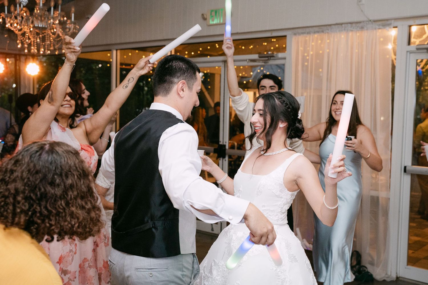 Bride and groom dancing with guests holding colorful glow sticks at a festive wedding reception.