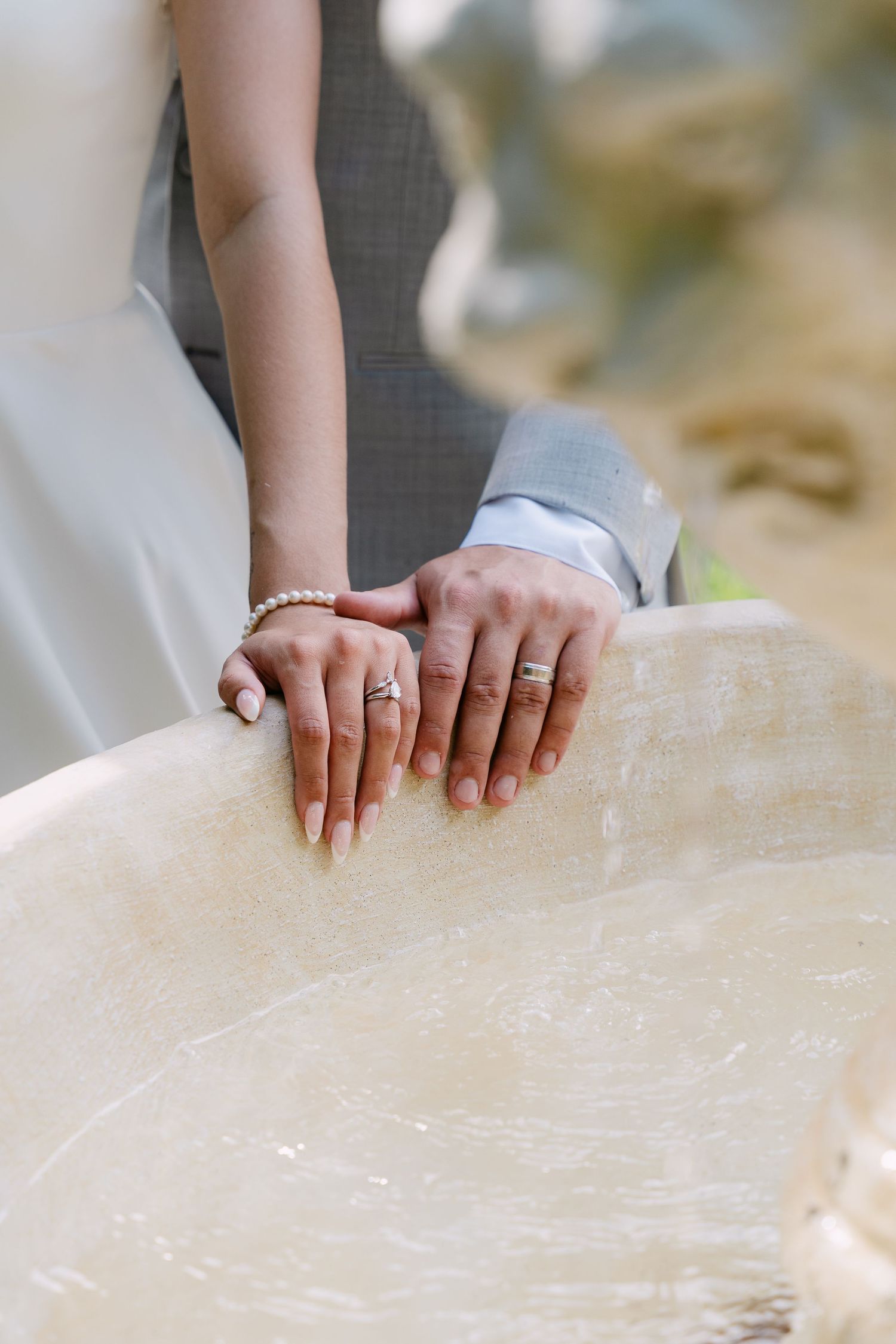 Newlywed couple resting hands on stone surface, showing wedding rings and pearl bracelet.