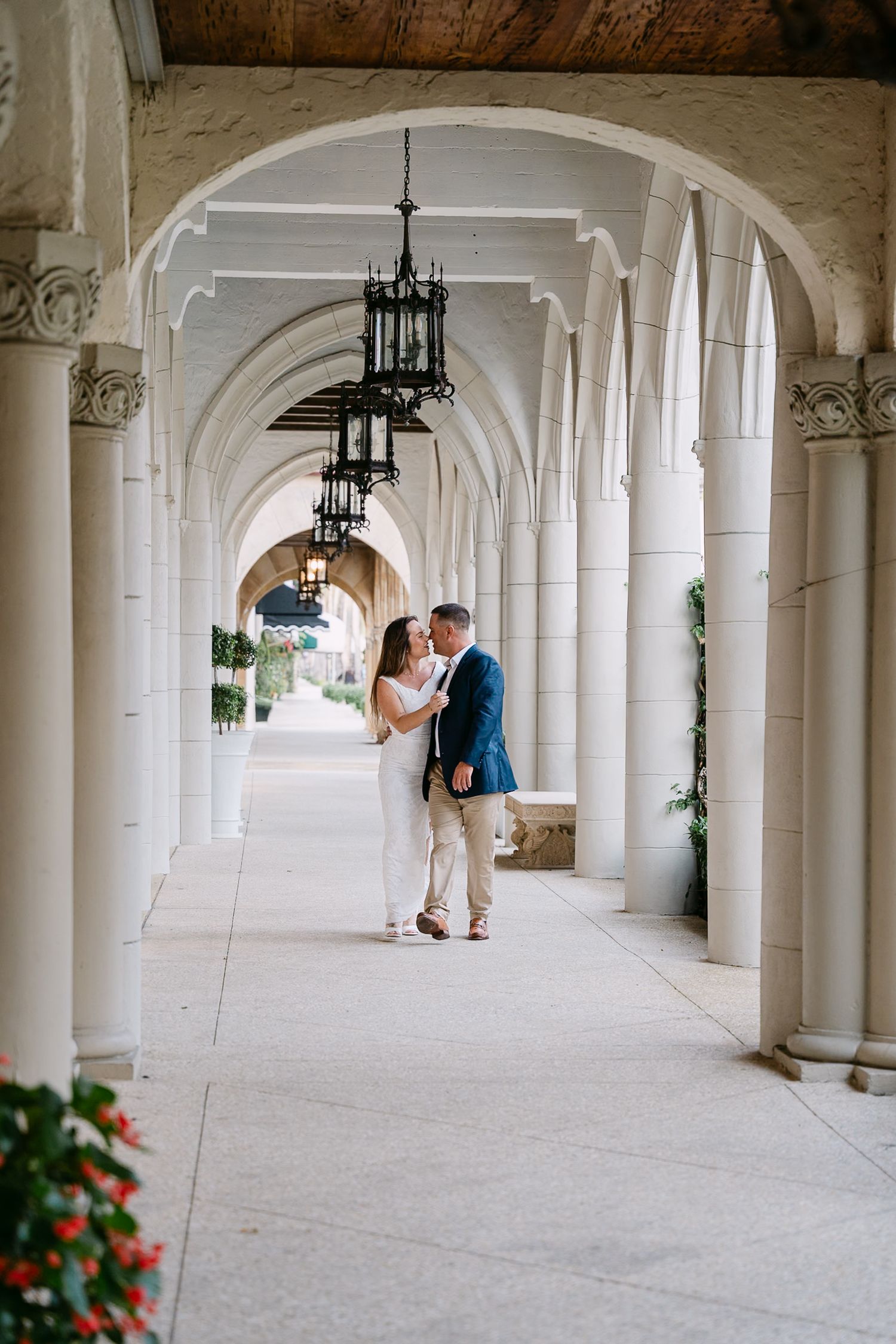 Engaged couple sharing a tender kiss in a stunning white arched colonnade with hanging lanterns.