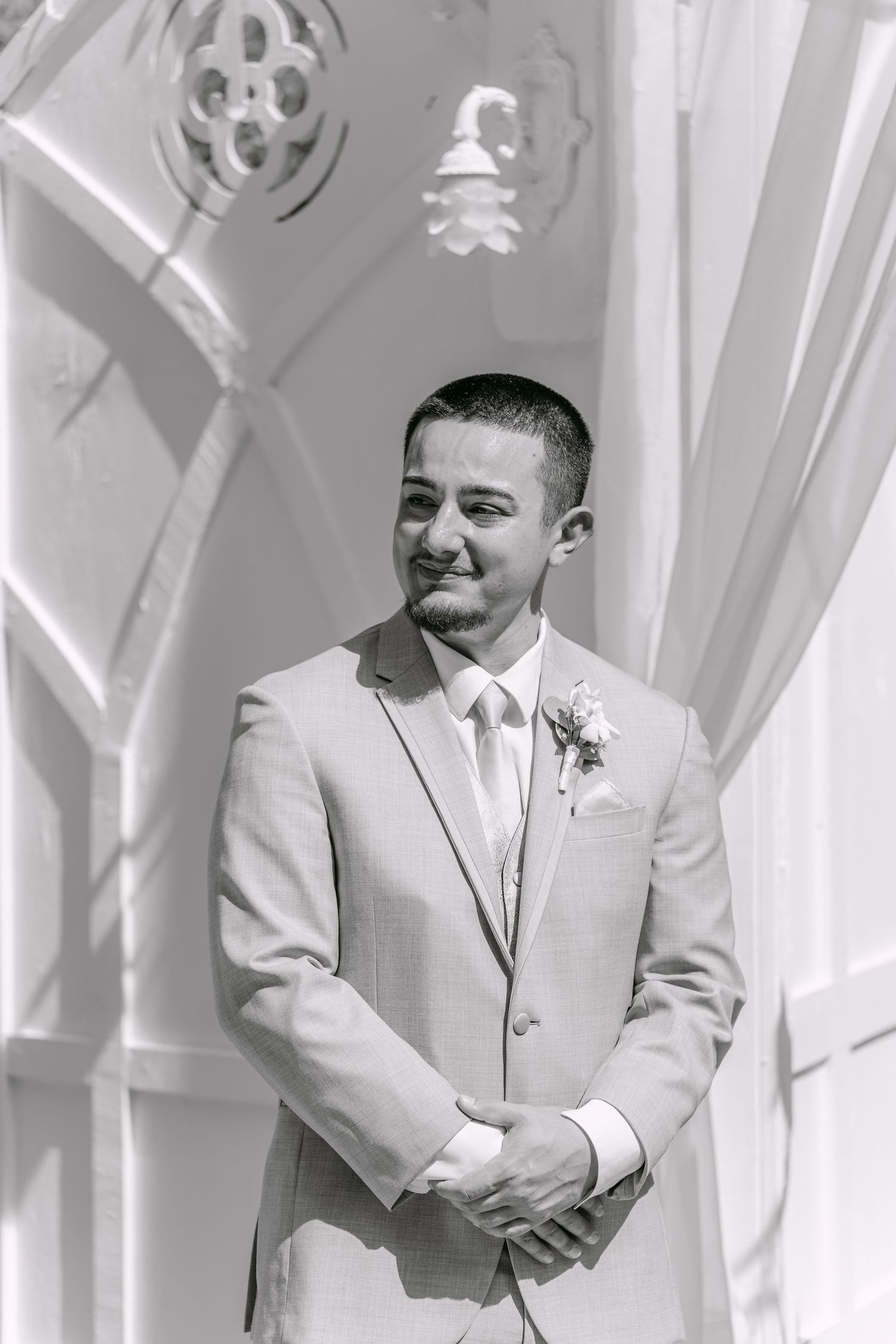 Black and white photo of a groom in a light suit with boutonniere, smiling at a wedding ceremony.