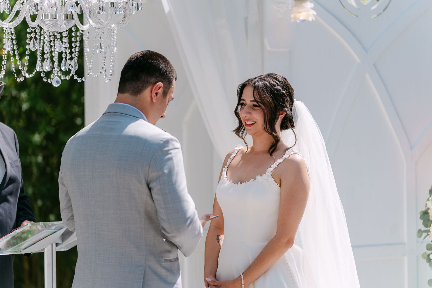 Bride in lace wedding gown smiling at groom during outdoor wedding ceremony under crystal chandelier.