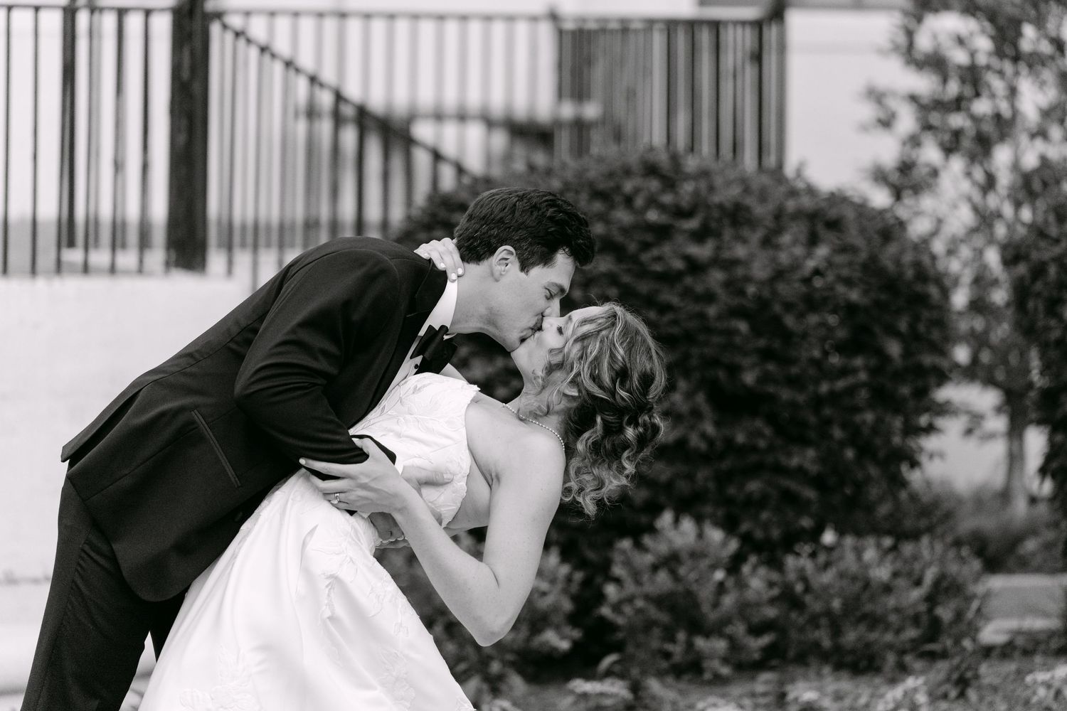 Elegant black and white wedding photo of groom dipping bride for a passionate kiss outdoors.