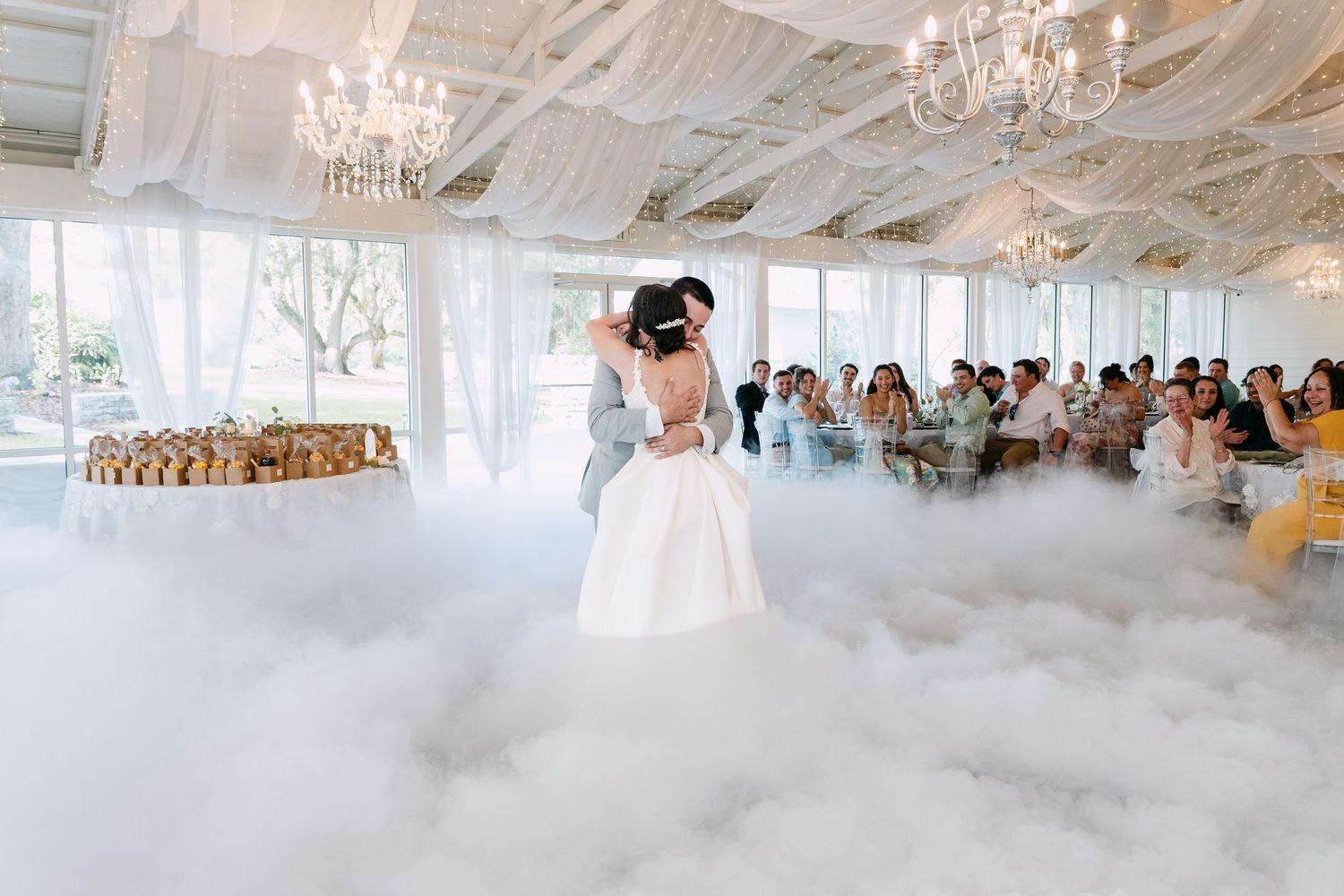 Bride and groom share first dance surrounded by dramatic fog on wedding dance floor with chandeliers and guests.