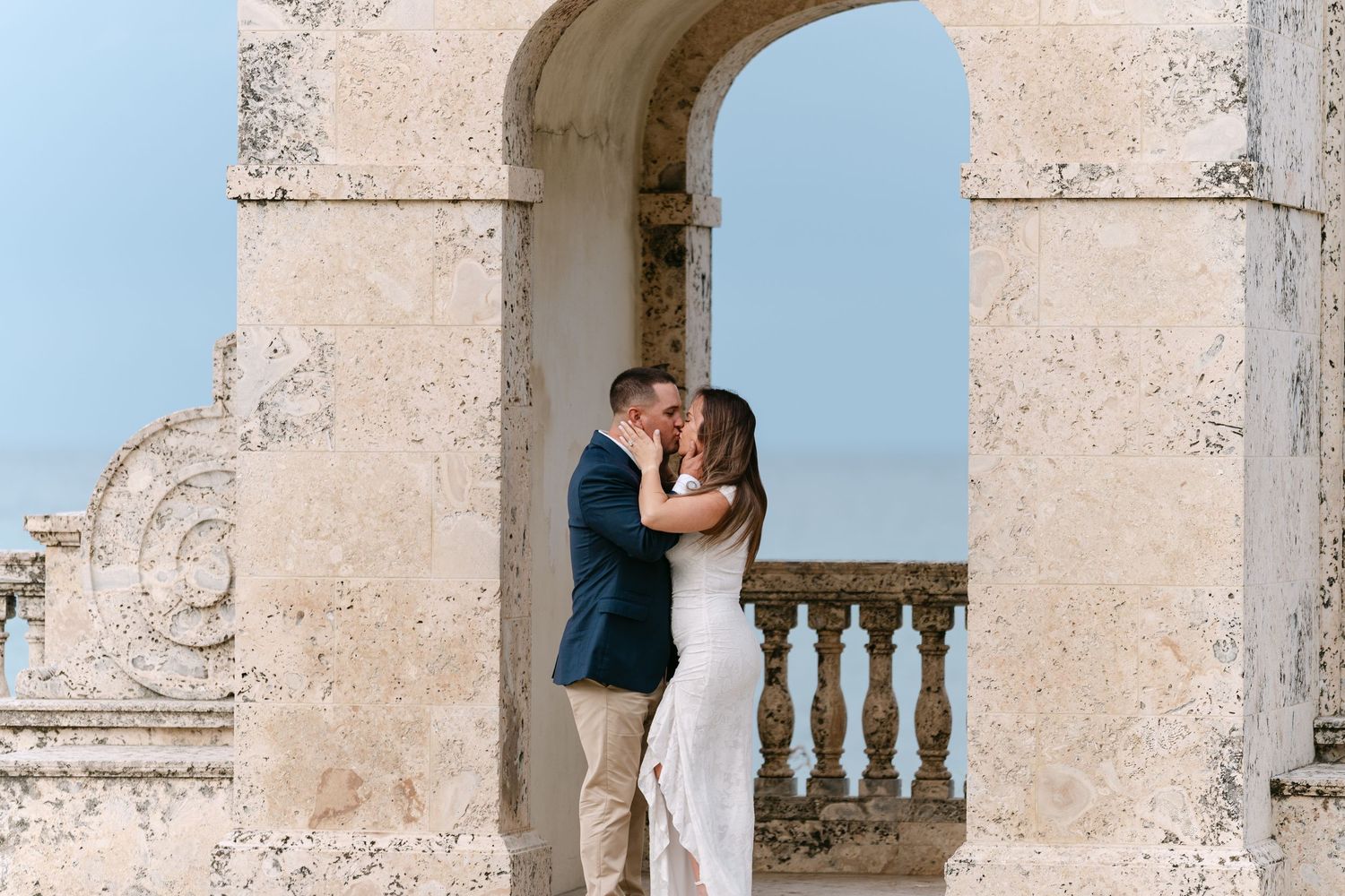Couple sharing a romantic kiss framed by a stone archway with ocean views at Vizcaya Museum.