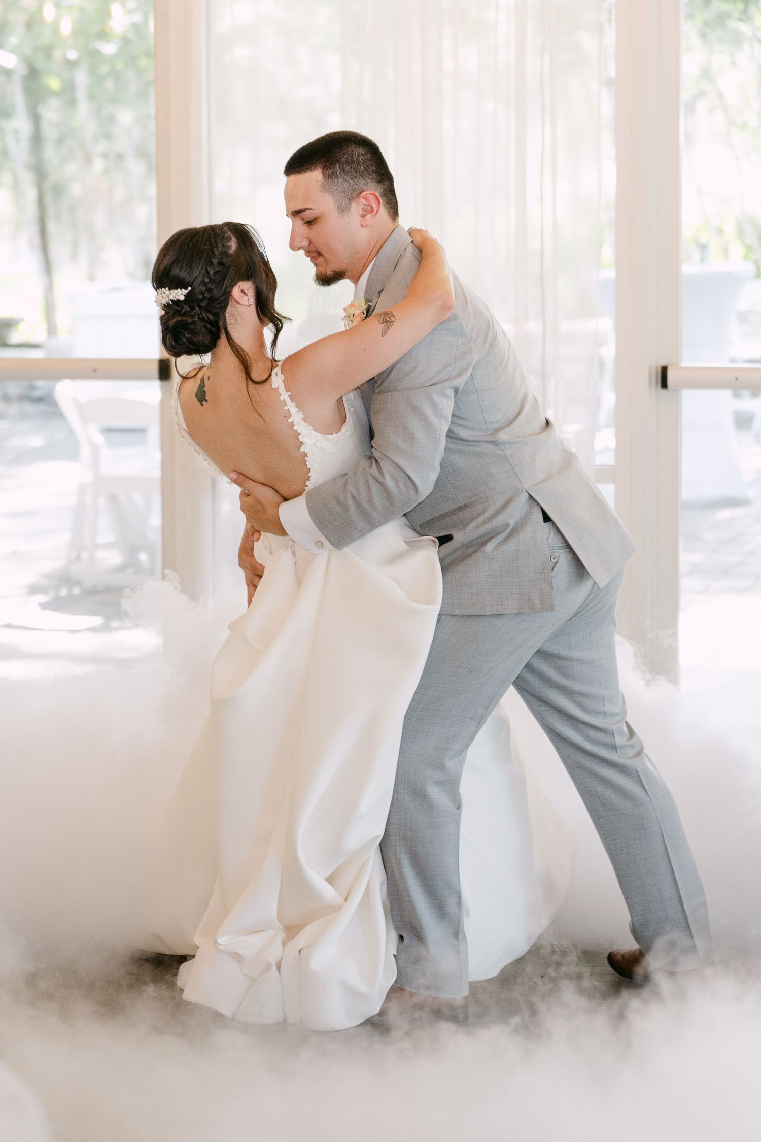 Groom dips bride in white gown on misty dance floor at wedding.