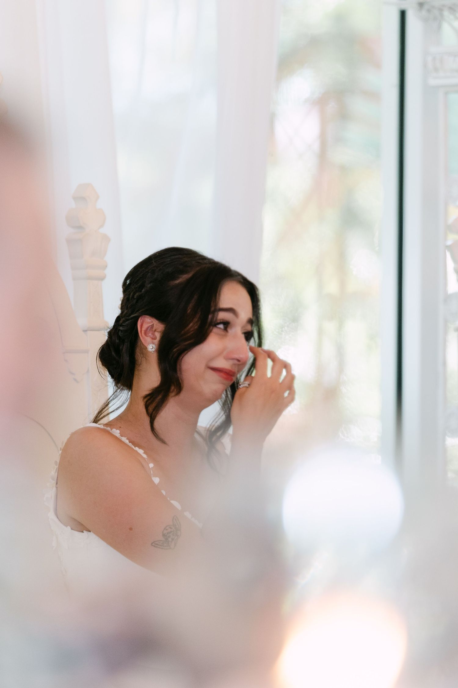 Emotional bride wiping tears, wearing white dress and pearl earrings, seated near bright window with soft natural light.