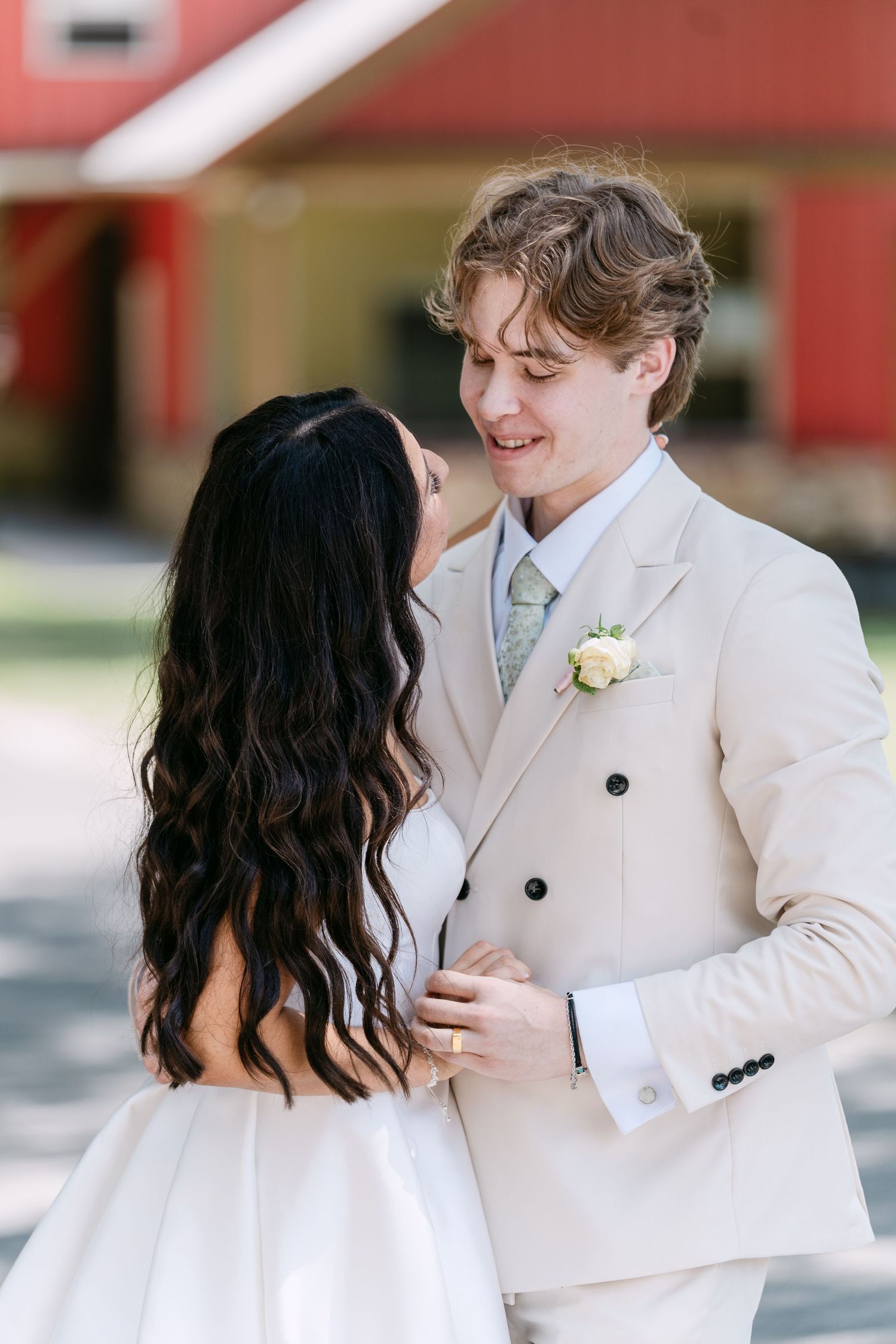 Groom in beige suit with boutonniere smiles at dark-haired bride in white dress during outdoor wedding ceremony.