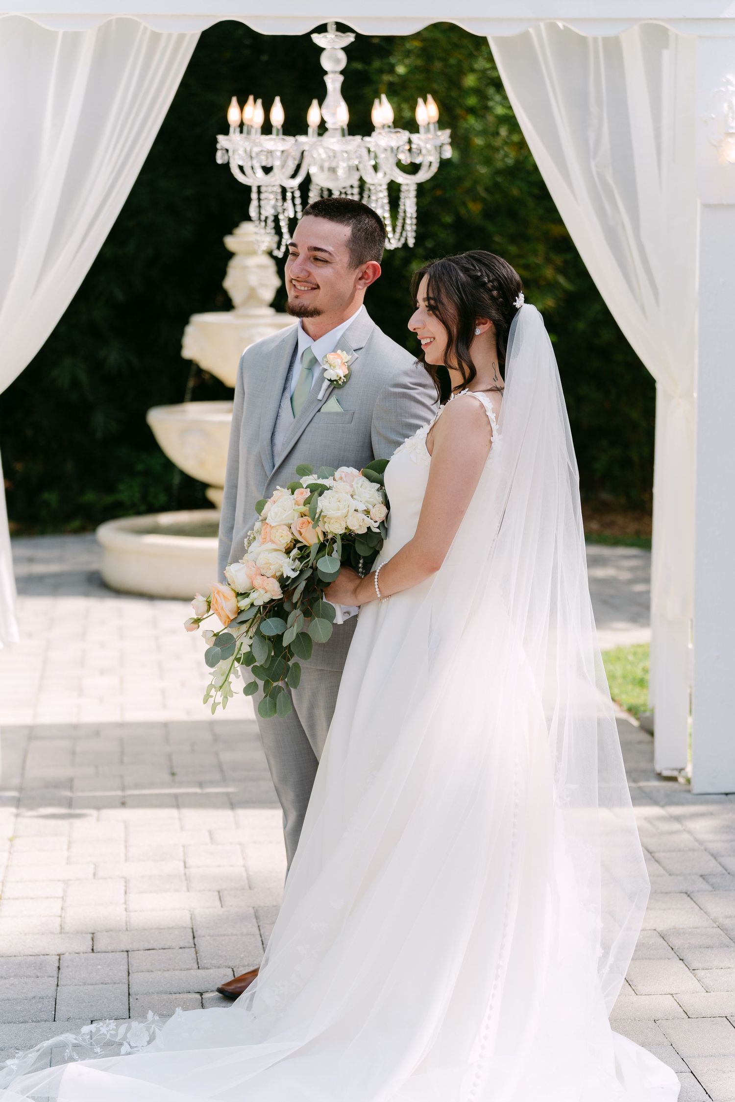Bride and groom share a romantic moment under a crystal chandelier arch with white draping and a fountain backdrop.